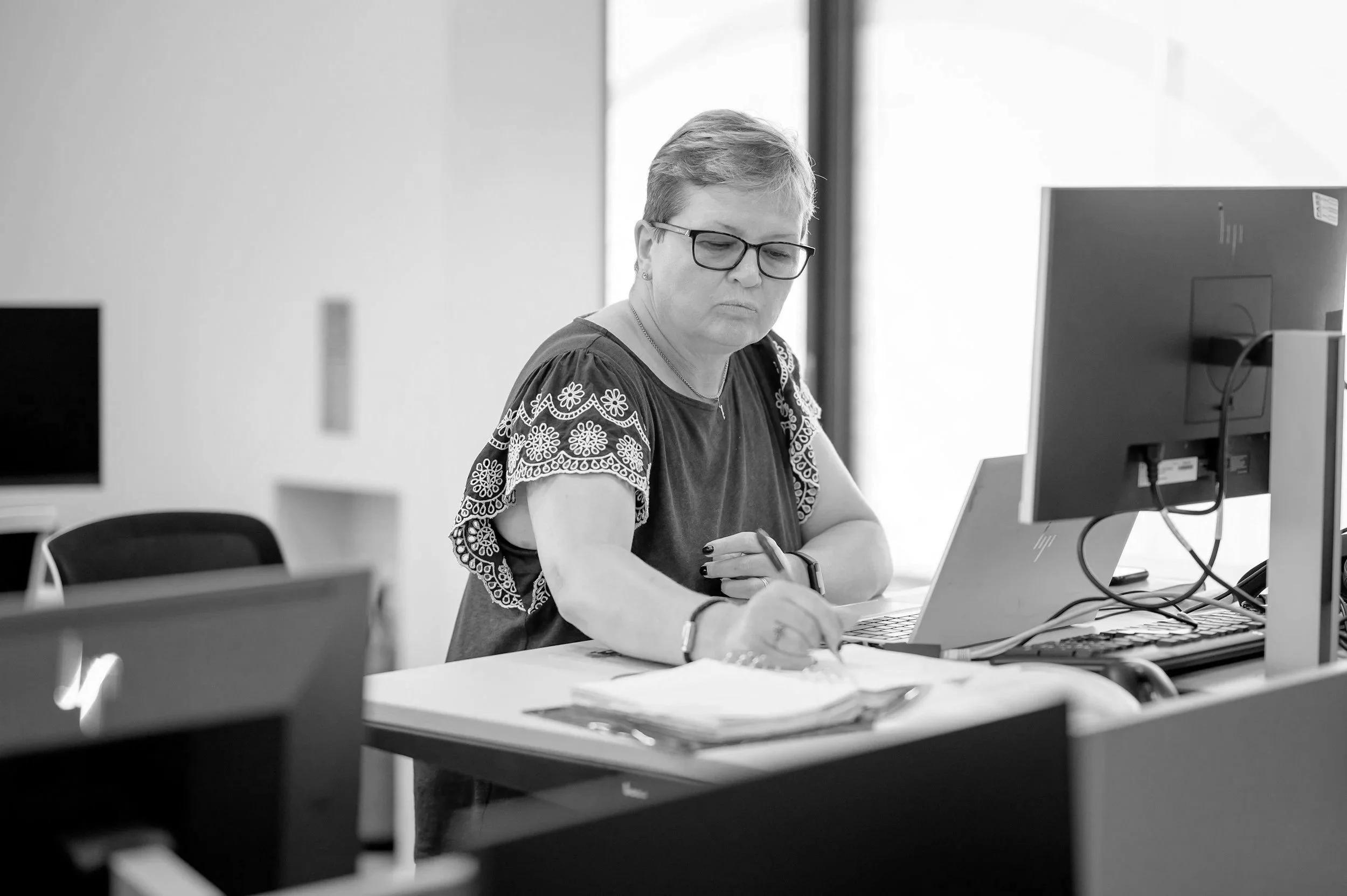 A woman working at her desk in an office, wearing glasses and a black top with floral embroidered sleeves, writing notes beside a computer monitor.