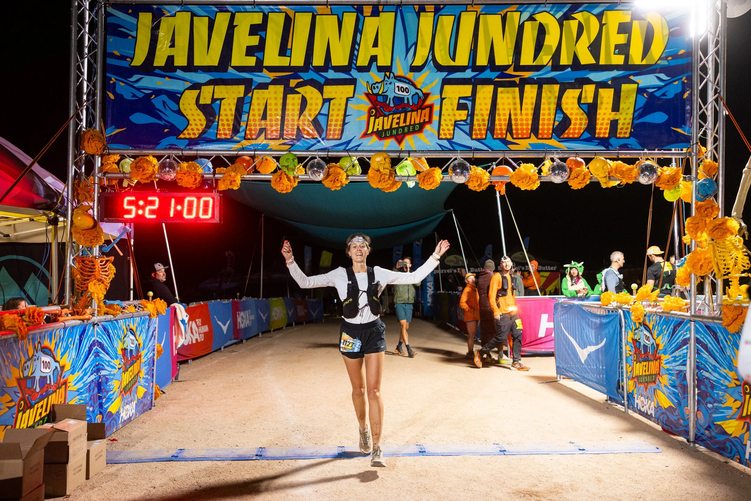 Runner crossing the finish line at the Javelina Jundred race during the night, with a large colorful banner overhead and a digital clock showing 5:21:08.