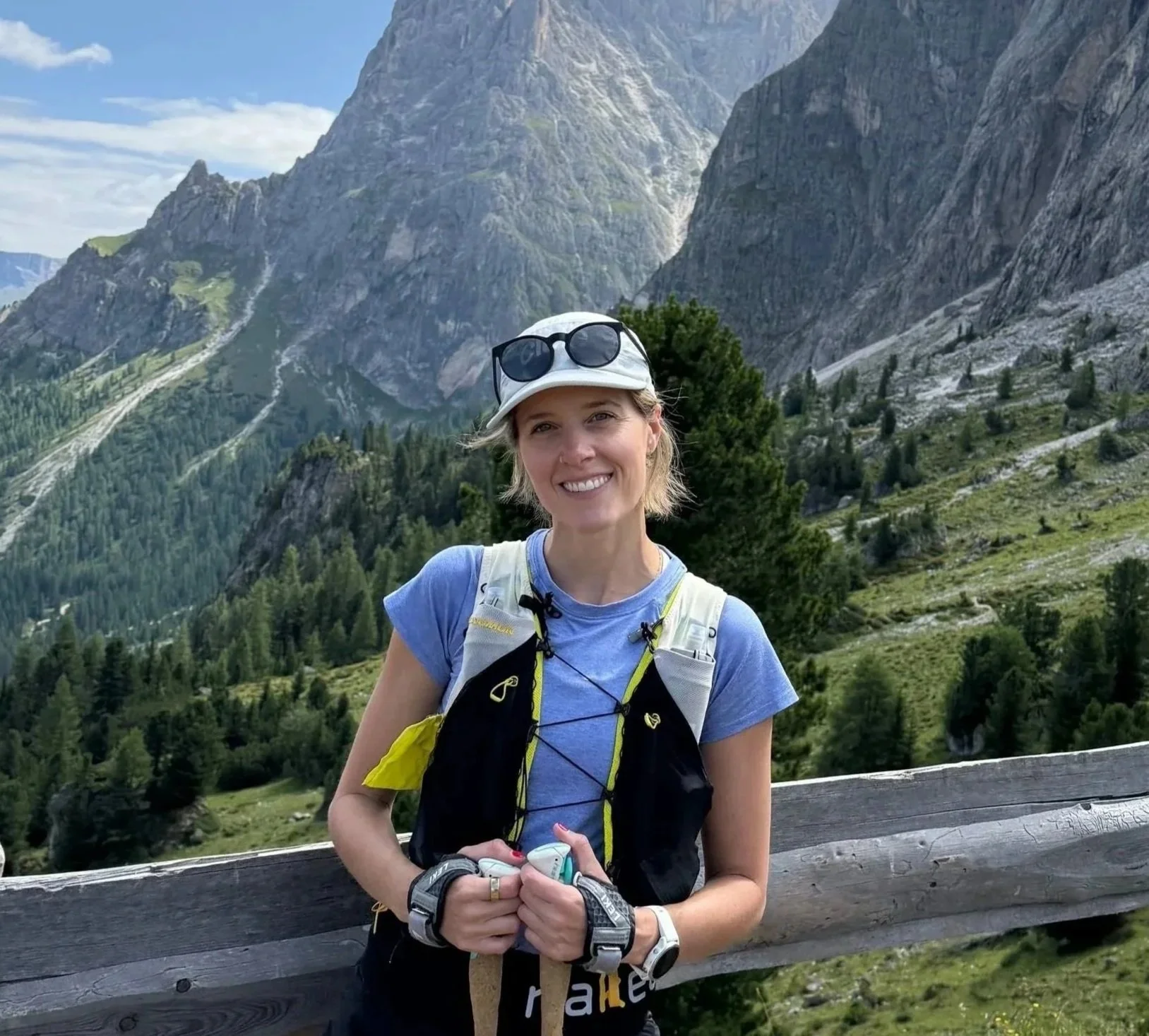 A woman standing outdoors on a mountain trail, smiling, with tall mountains and green trees in the background, wearing a blue shirt, white hat with sunglasses, and a hydration vest.
