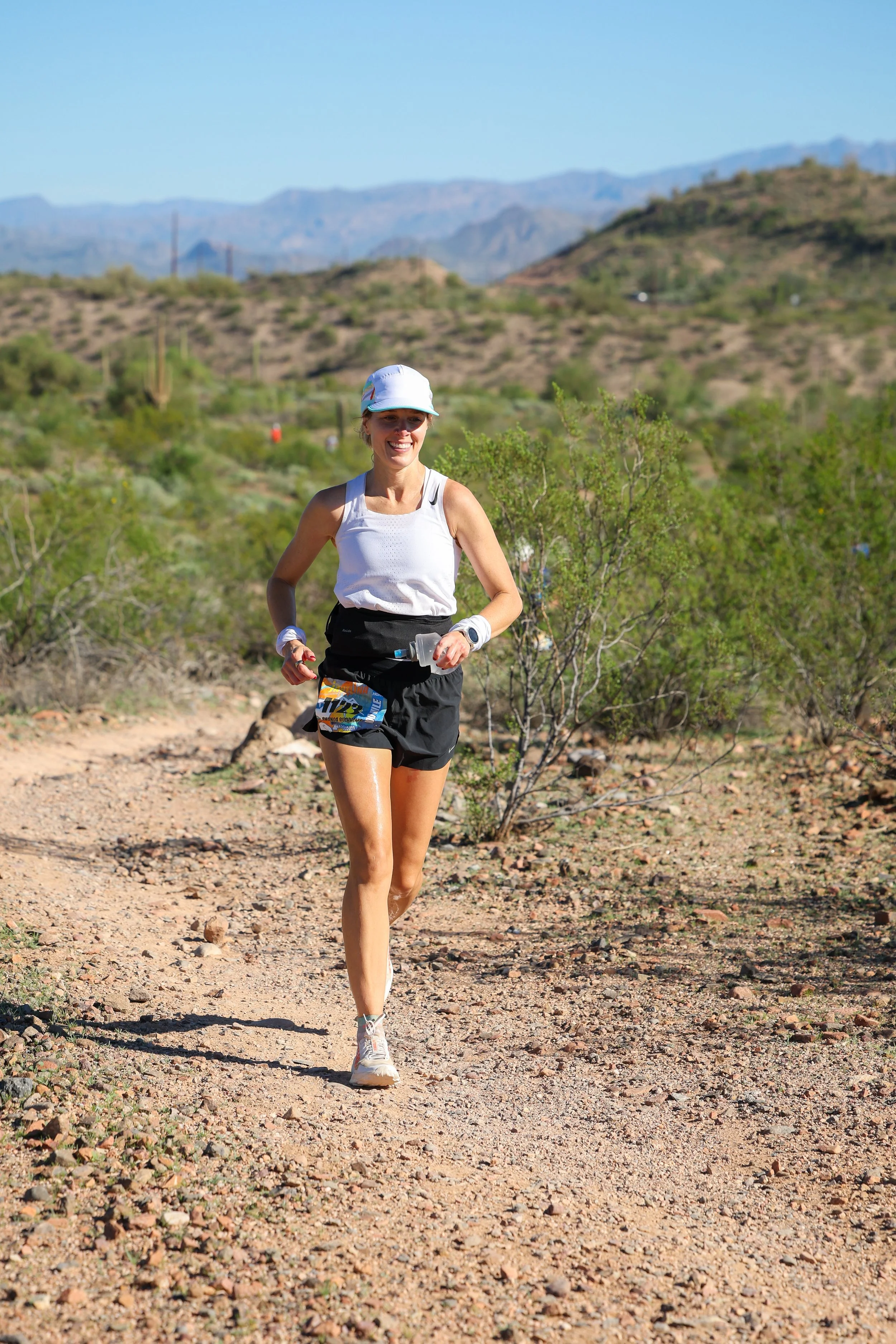 A woman running on a dirt trail in a desert landscape with mountains in the background, wearing a white tank top, black shorts, a cap, and a race bib.