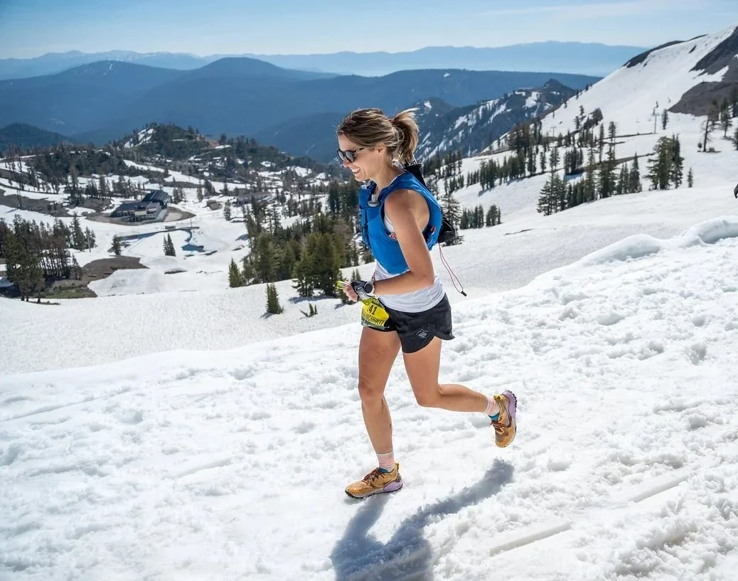 A woman running on snow-covered mountains wearing athletic clothes, sunglasses, and a backpack.