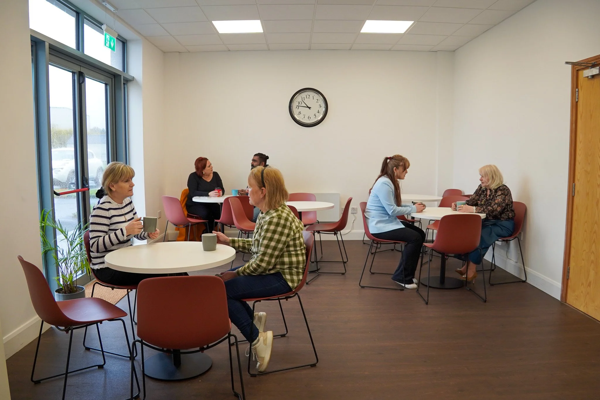 Six women sitting at separate white tables in a room with white walls, large windows, and a clock showing 9:10. Four women are seated at two tables, having conversations and holding mugs. The room has a wooden floor and a door on the right. One woman wears a striped sweater, another a checked shirt, and others in casual clothing.