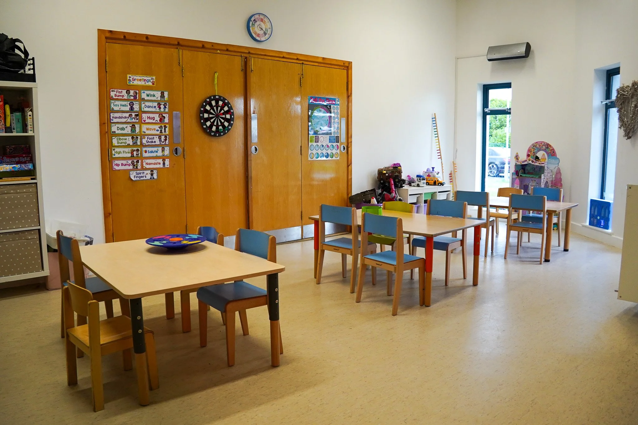 Empty classroom with colorful child-sized tables and chairs, a wall decorated with educational posters, a dartboard, toys, and large windows letting in natural light.