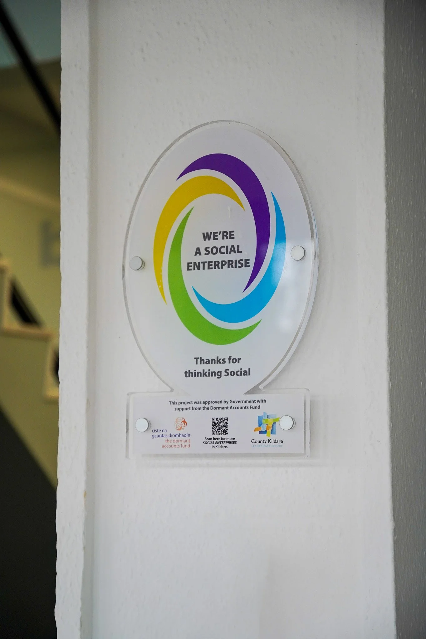 Sign on a white wall that reads, "We're a social enterprise," with colorful circular graphics around it, and additional text below that mentions support from the Dormant Accounts Fund, County Kildare, and social enterprise information.