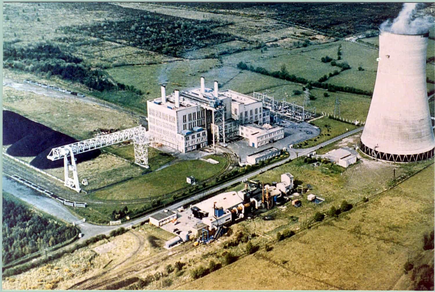 Aerial view of a nuclear power plant with a large cooling tower, a reactor building, and surrounding fields.