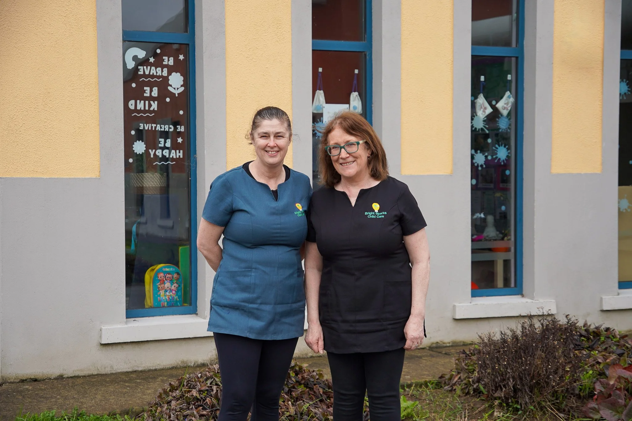 Two women standing outside a childcare center with yellow and gray walls and large window, smiling at the camera.