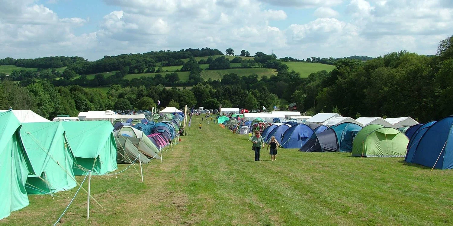 Tent camp area on a grassy field with many colorful tents, a few people walking along a path, and rolling green hills in the background under a partly cloudy sky.