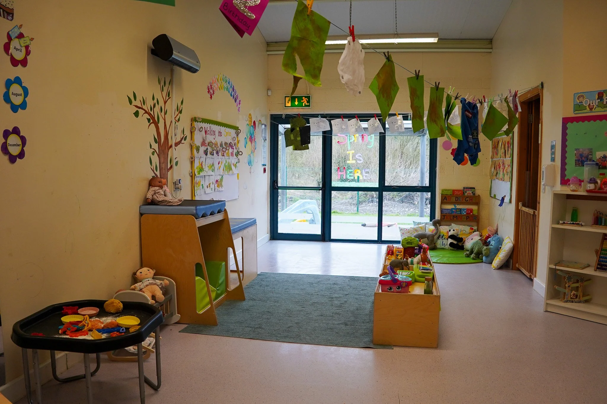 Colorful preschool or daycare classroom with toys, stuffed animals, and children's decorations. A sign reads "Spring is Here" in colorful letters.