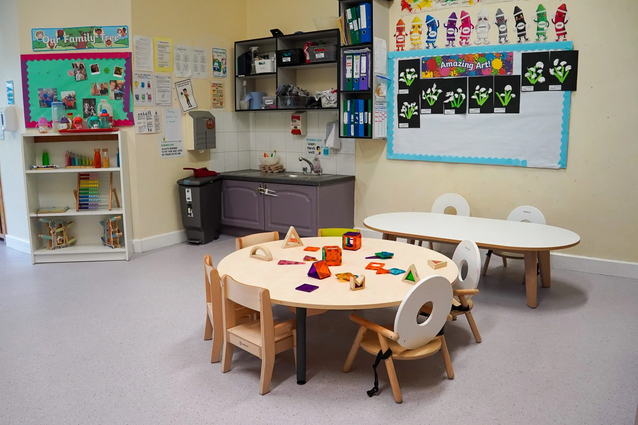 A classroom corner with educational toys on a round table, small chairs, a blackboard with children's artwork, shelves with toys and supplies, a kitchenette with a sink, a bulletin board, and colorful decorations on the wall.