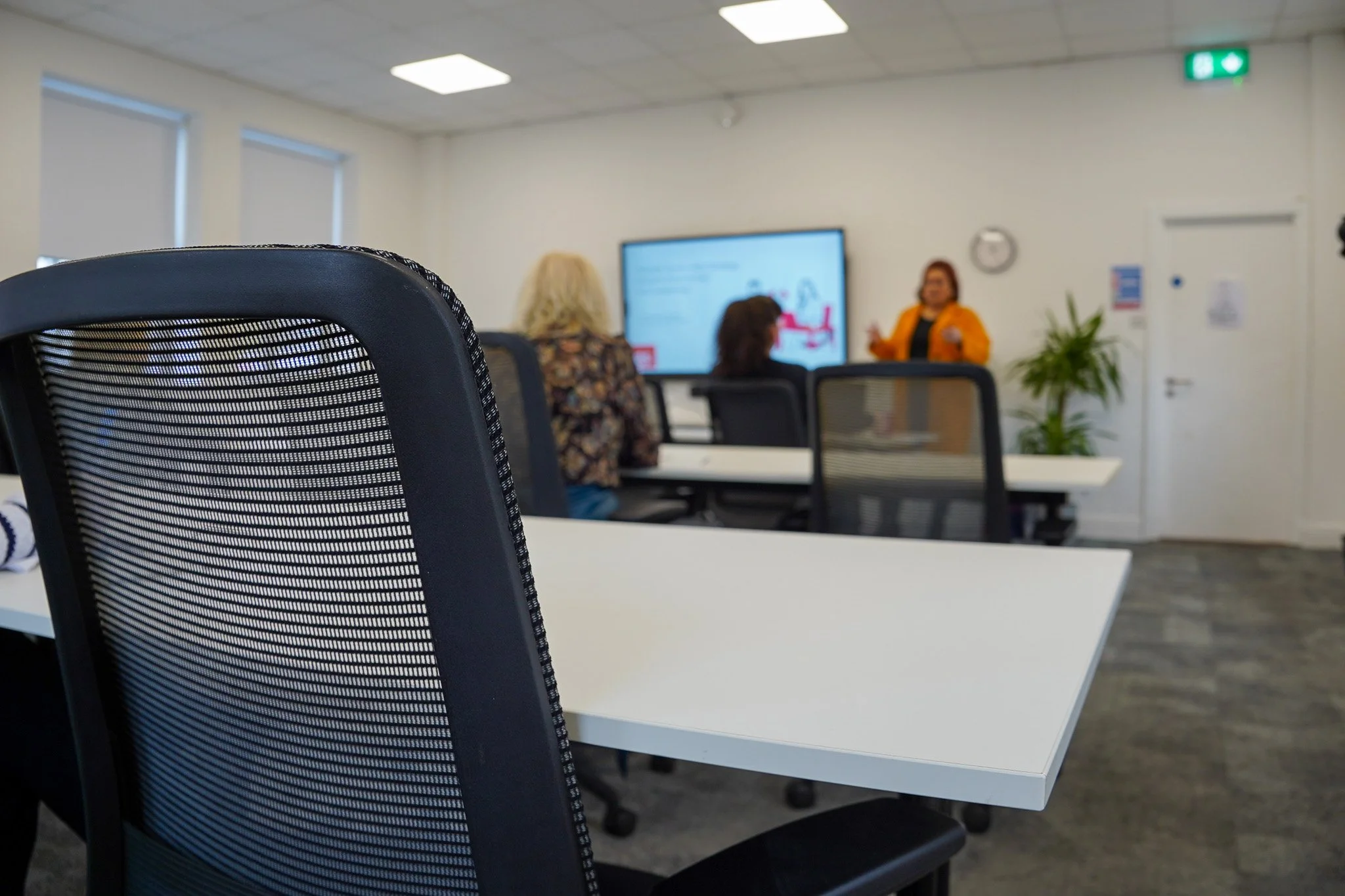 A classroom or meeting room with empty black mesh chairs and a presenter at the front near a large screen. Three people are seated and facing the presenter.