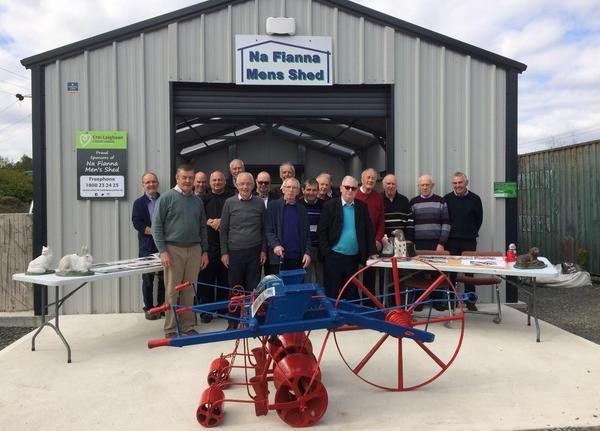 Group of men standing in front of a metal building labeled 'Na Fianna Mens Shed', with a table on each side and an old-fashioned red farm plow in the foreground.