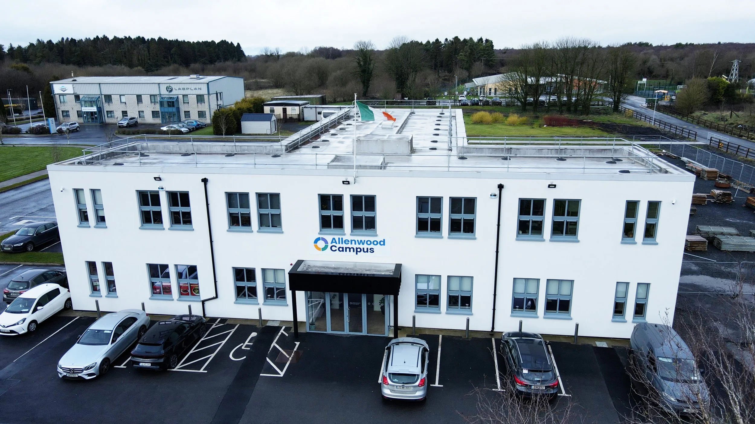 Aerial view of a white commercial building labeled 'Allenwood Campus' with parking lot and cars in front, surrounded by trees and other industrial buildings.