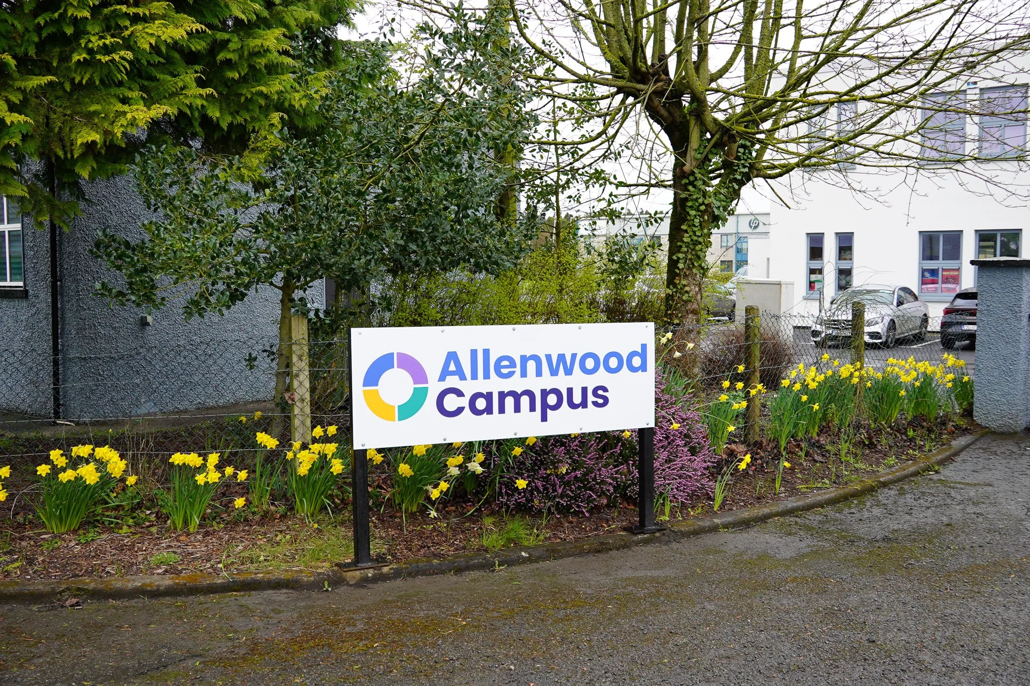 Sign reading 'Allenwood Campus' in front of a flower bed with yellow flowers, surrounded by trees and a chain-link fence, near parked cars and a building.