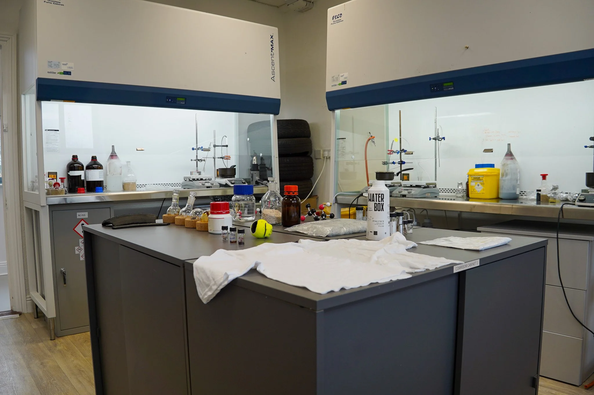 A scientific lab with two fume hoods, a central table with laboratory supplies, chemicals, and glassware, and a whiteboard in the background.