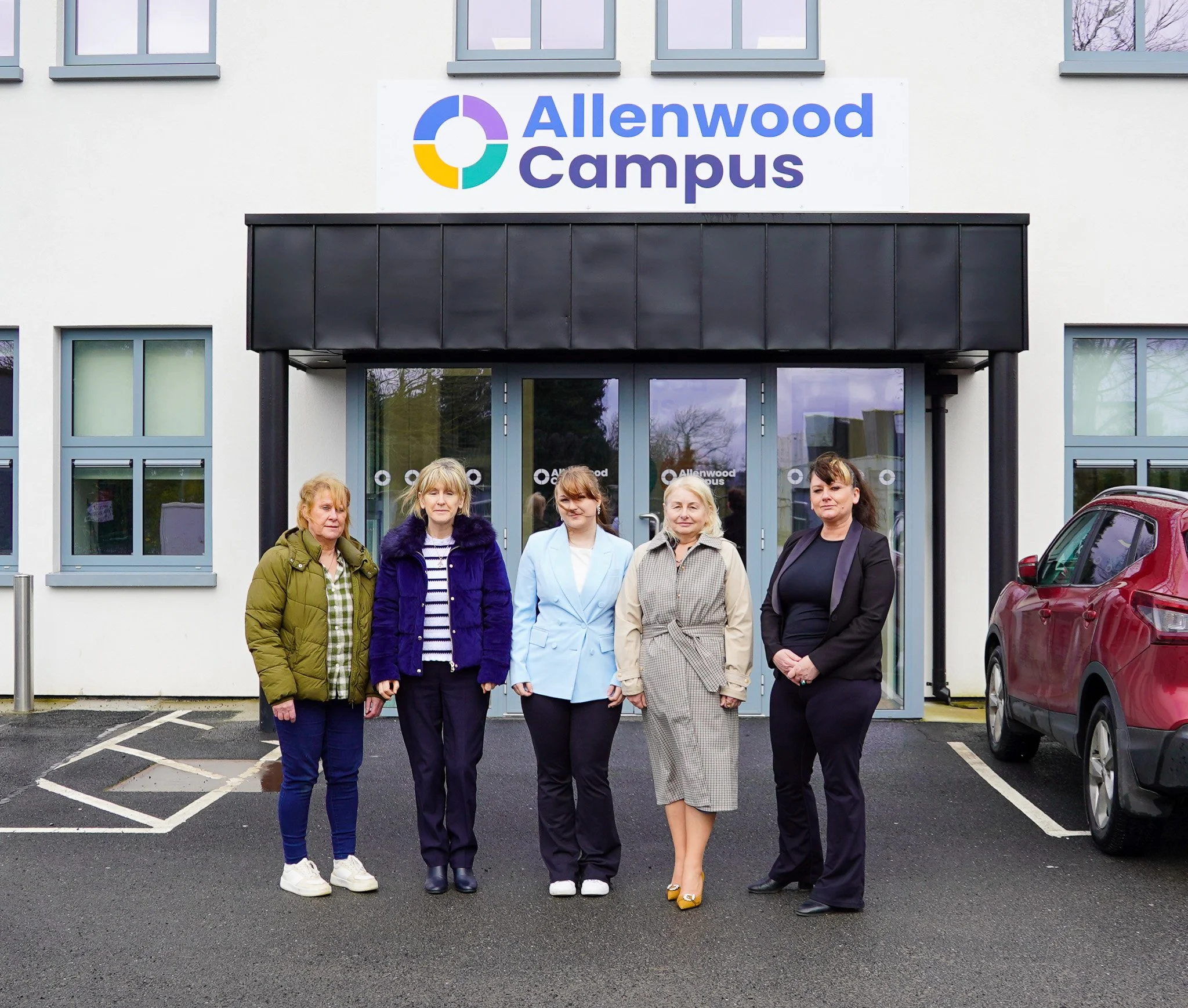 Five women standing in front of a building with a sign that reads 'Allenwood Campus'; there are parking spaces and a red car visible on the right.