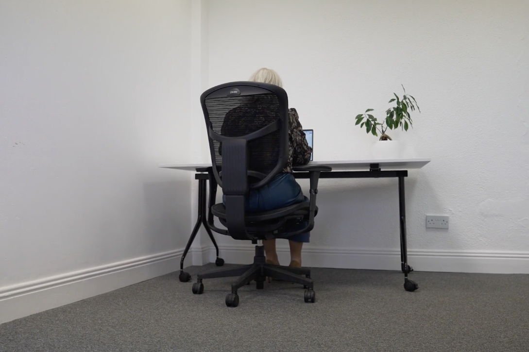 A person sitting at a desk working on a laptop in a minimal office, with a potted plant on the desk, white walls, and gray carpet.