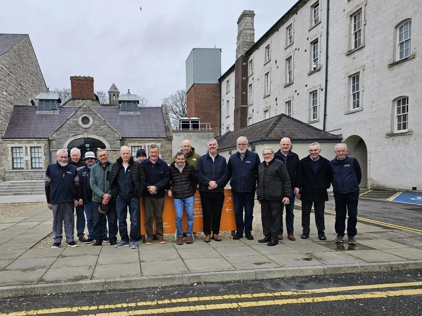 A group of people, mostly older men, standing outdoors on a paved area in front of historic buildings, posing for a photo.