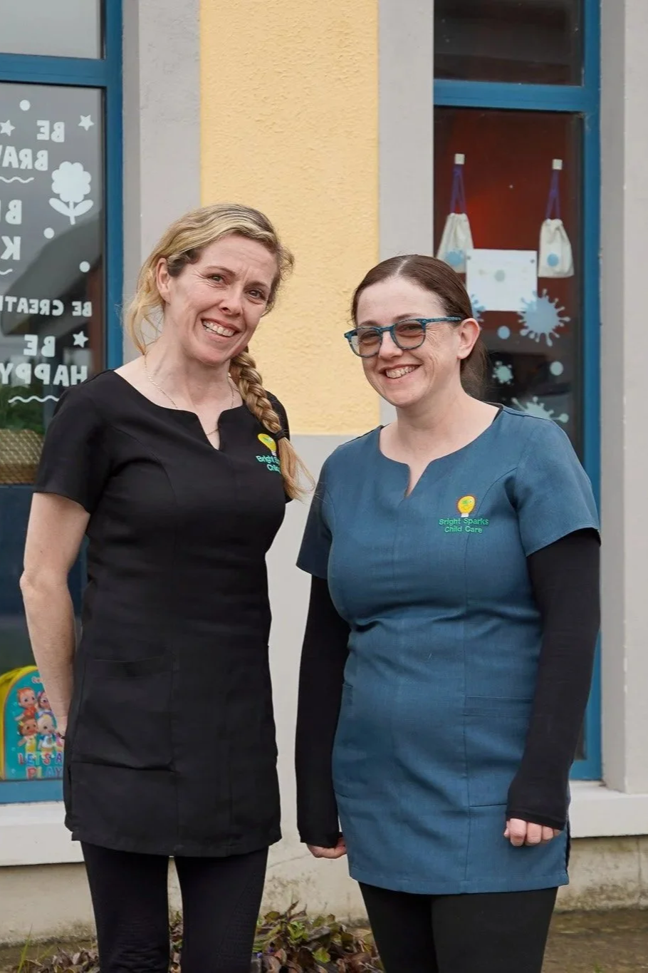 Two women standing outside a childcare center, smiling for the photo. Both are wearing uniforms with the logo of Bright Sparks Child Care.