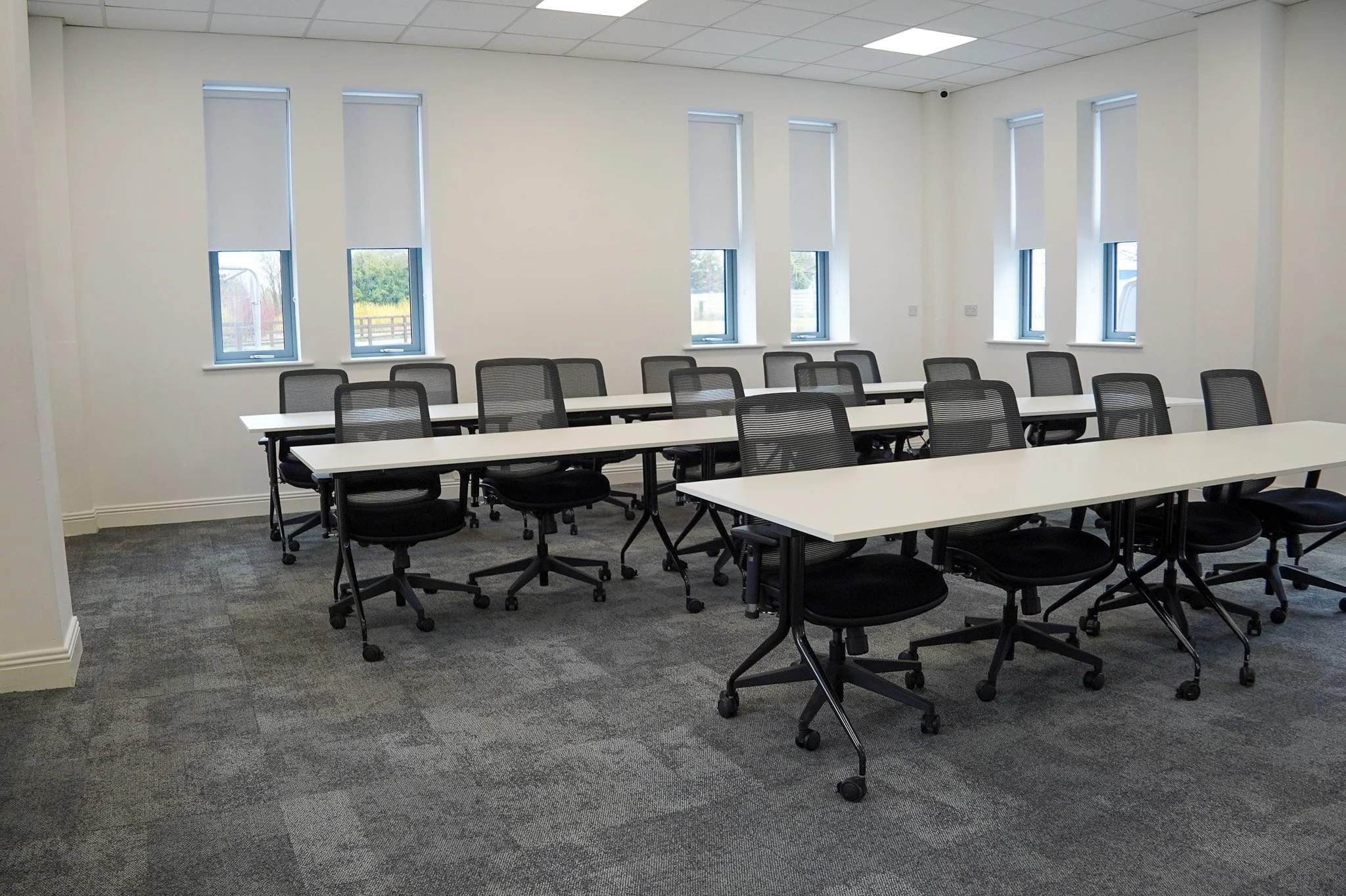 An empty conference room with white tables and black mesh office chairs, illuminated by natural light through five windows with white blinds.