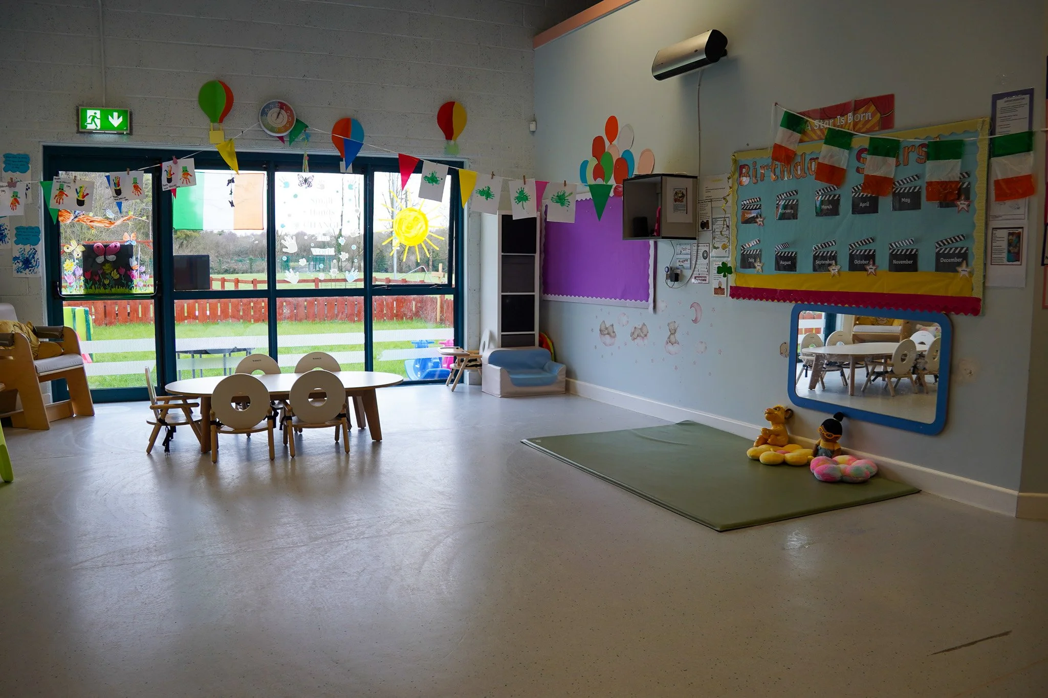 Empty classroom or playroom with colorful decorations, furniture, and toys, large windows with outdoor view, and educational materials on walls.