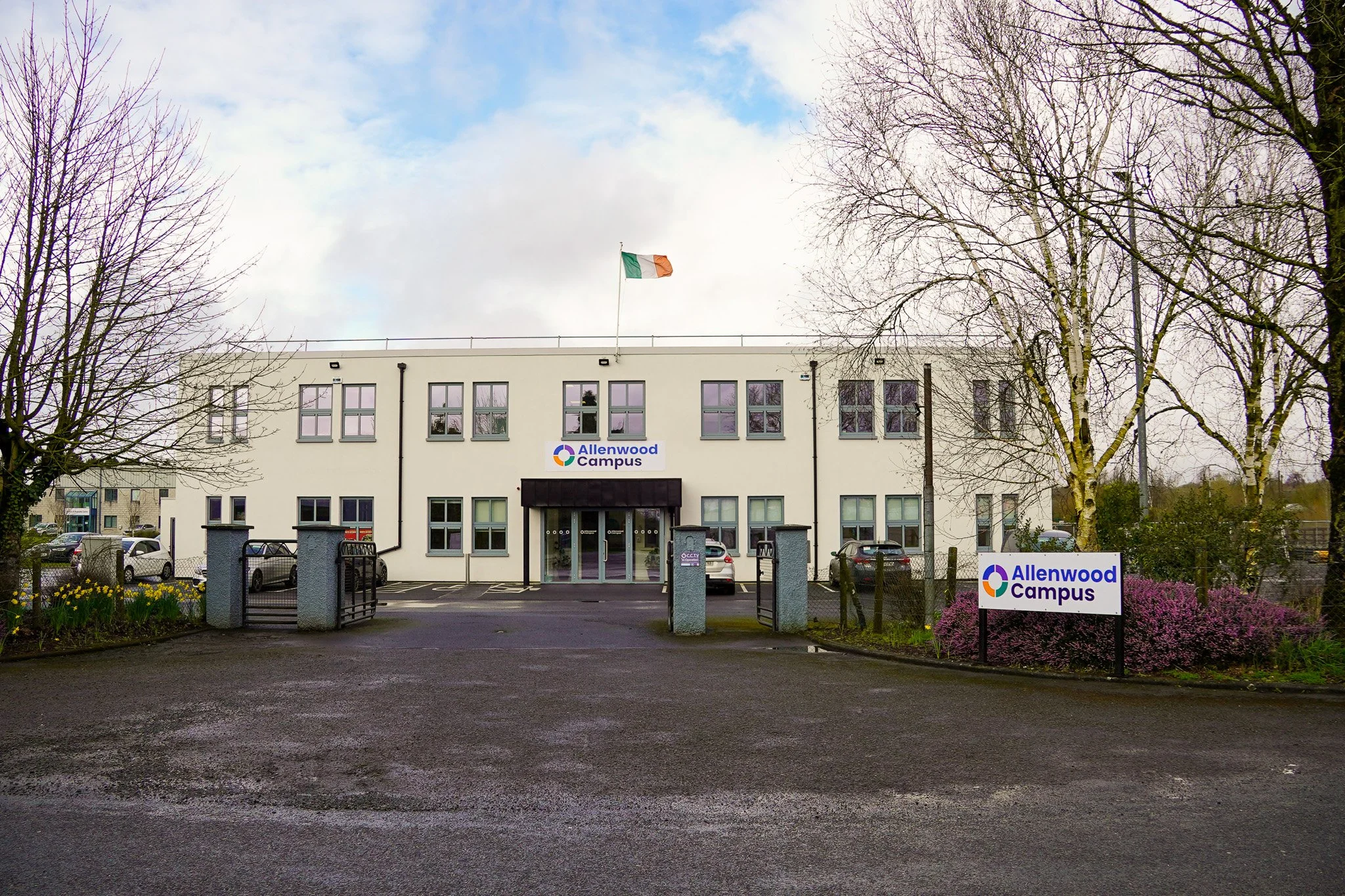 Front view of a white building with a sign that reads 'Allenwood Campus,' with an Irish flag on top, surrounded by trees and parked cars.
