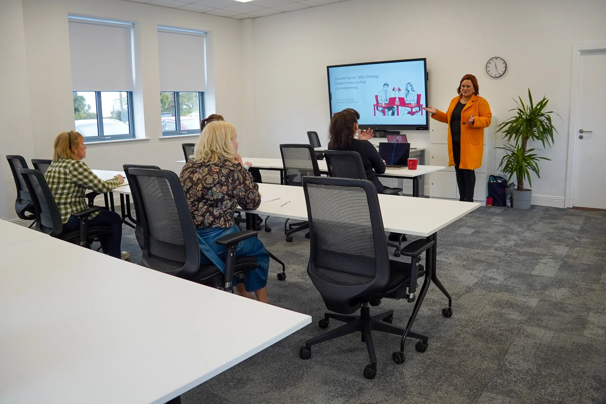 A woman giving a presentation to four women seated at tables in a conference room, with a large monitor displaying a slide titled 'Growth Sprint: Why Strategy Doesn't Have to Feel Overwhelming'.
