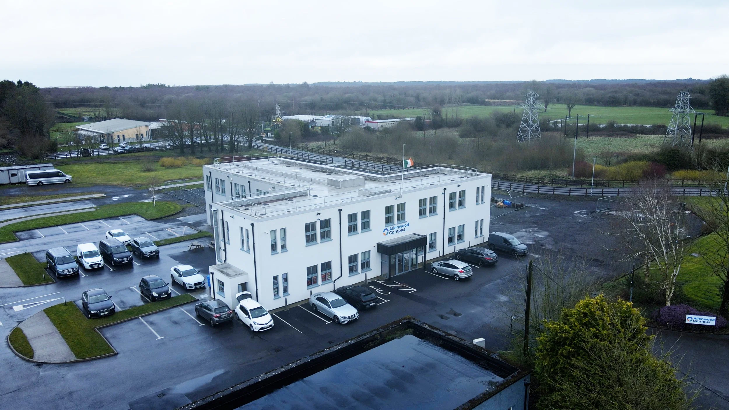 An aerial view of a white office building labeled 'Allenwood Campus' with a parking lot filled with cars, surrounded by greenery and open fields under a cloudy sky.