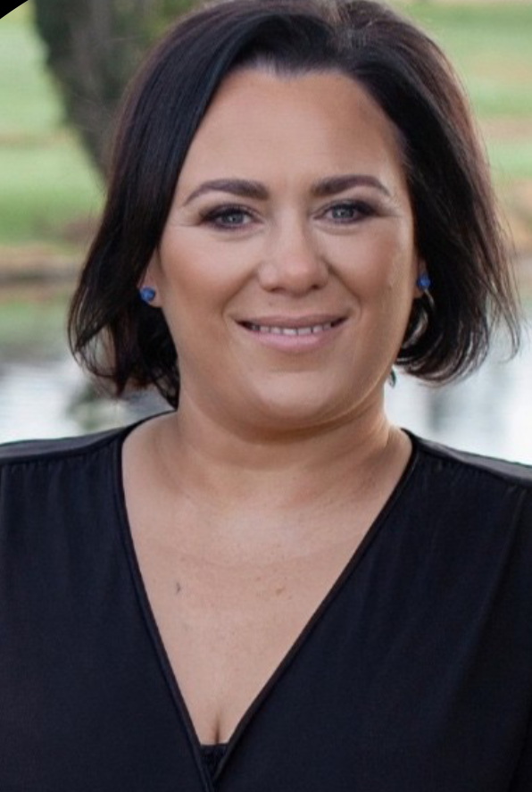 A woman with short dark hair, wearing a black top and blue earrings, standing outdoors near a body of water, smiling at the camera.