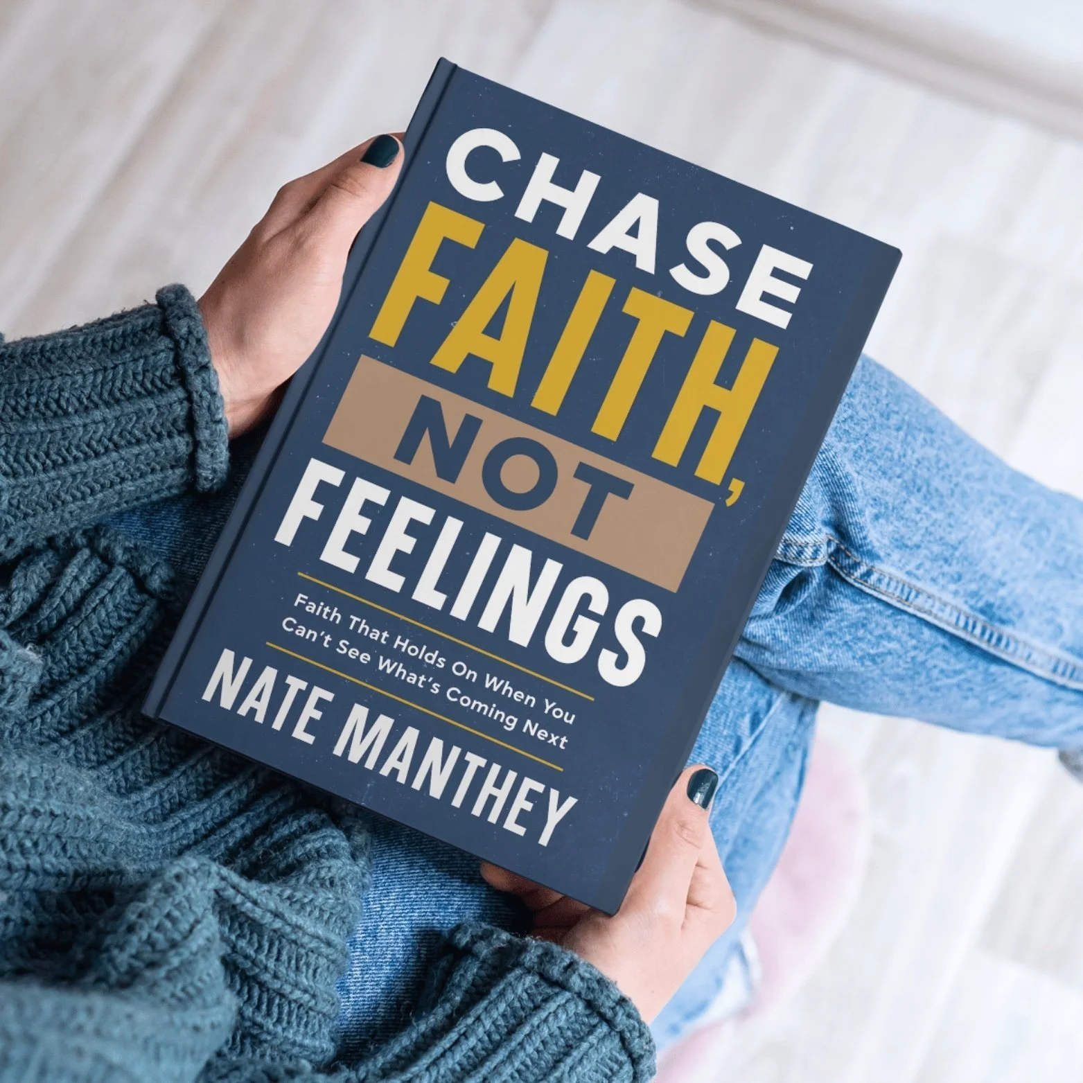 A person holding a book titled 'Chase Faith, Not Feelings' by Nate Manthey. The person is wearing a blue knitted sweater and blue jeans, sitting with one leg crossed over the other.