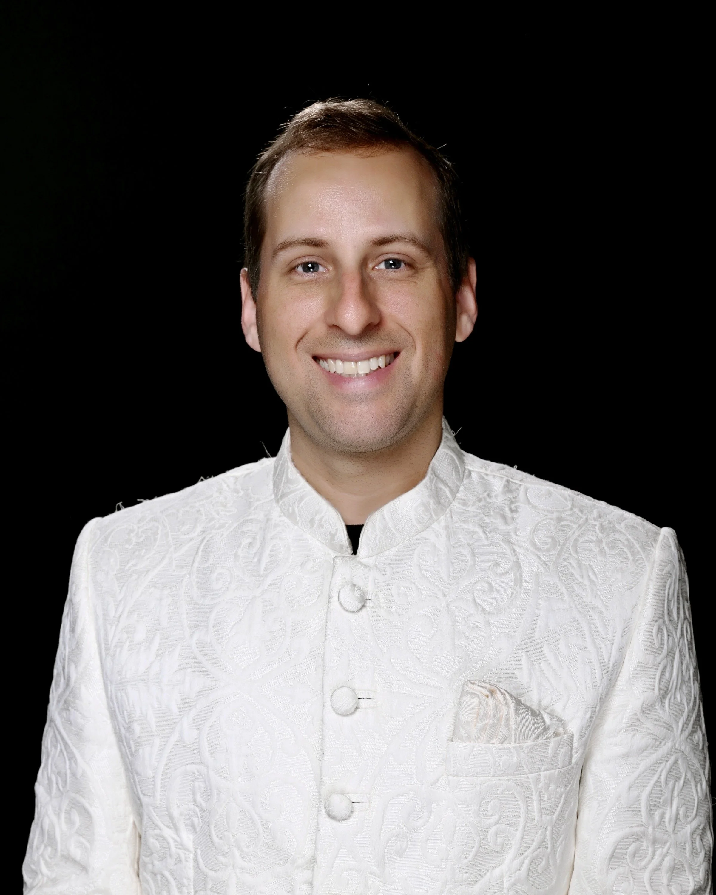 A smiling man wearing a white traditional Indian sherwani with embroidery, standing against a black background.
