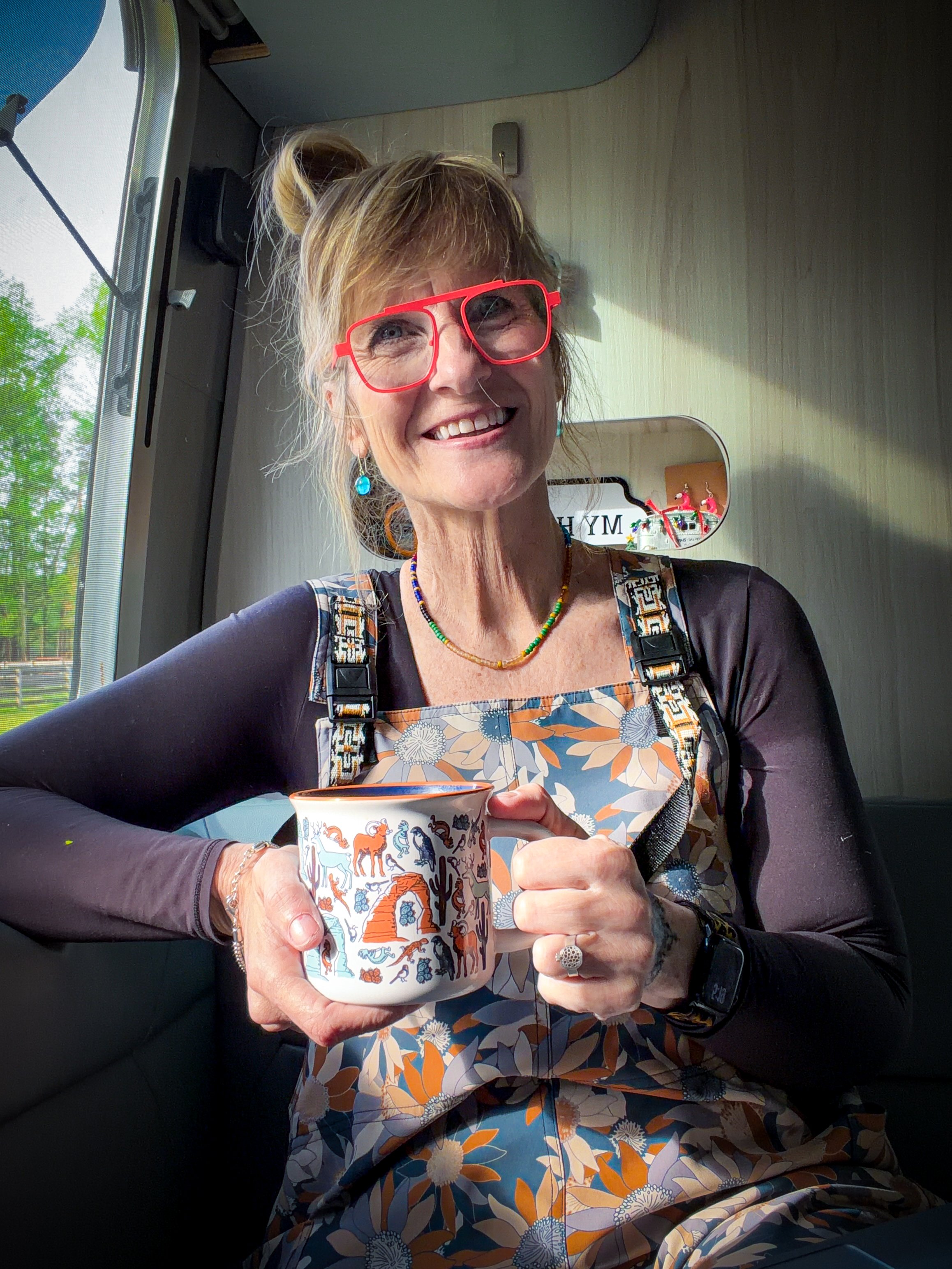 Pamela sitting insider her camper holding a mug of coffee with an illustrated design from Moab National Park. She is wearing orange glasses, floral overalls and a black long-sleeved shirt. She's smiling with her blonde hair in a loose bun.
