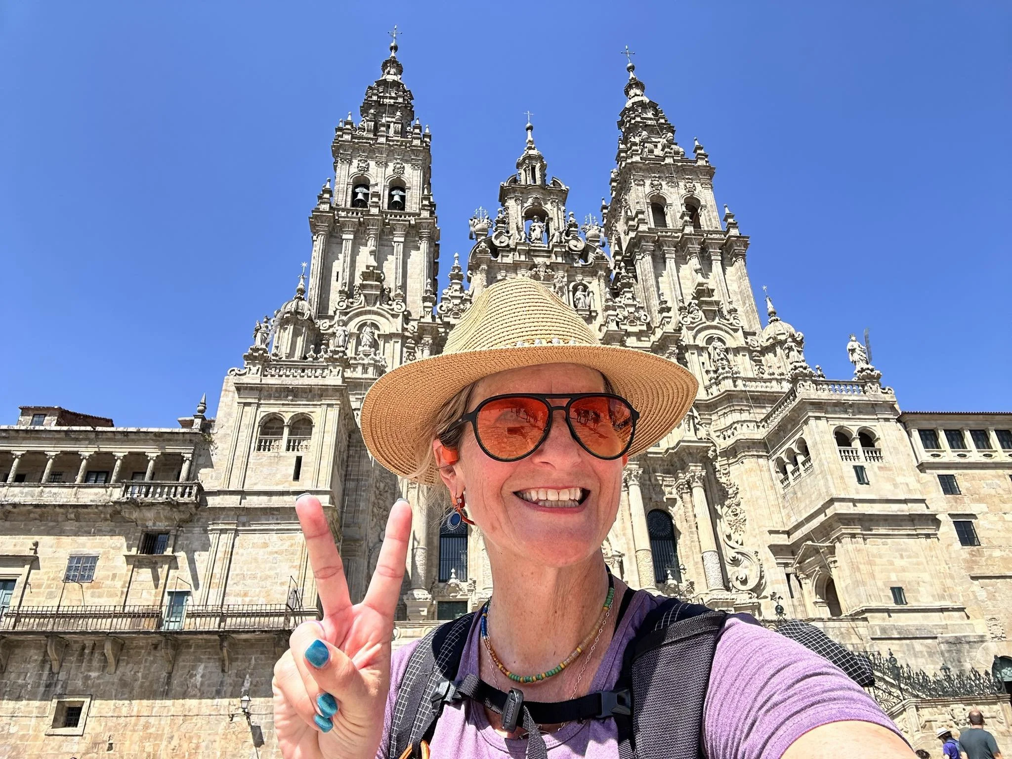 pamela with purple shirt, backpack, straw hat, sunglasses standing in front of the cathedral in the square at of Santiago De Compestela Spain on a Blue Sky Sunny Day. She signals 2 with her finger for her second complete camino.