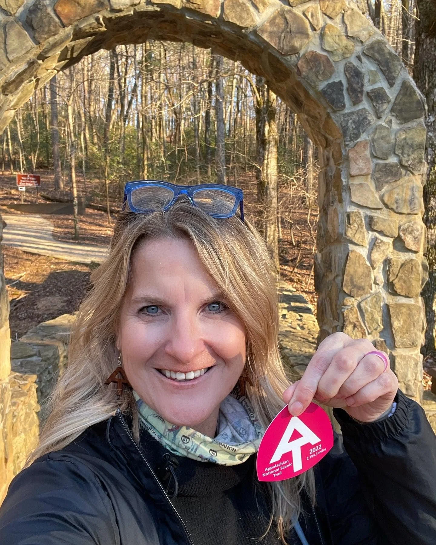 Pamela with blue glasses on her head, black pullover, neck buff, holding an AT 2022 red tag under the Arch at Amicalola Falls State Park in Georgia which is also the Southern Terminus