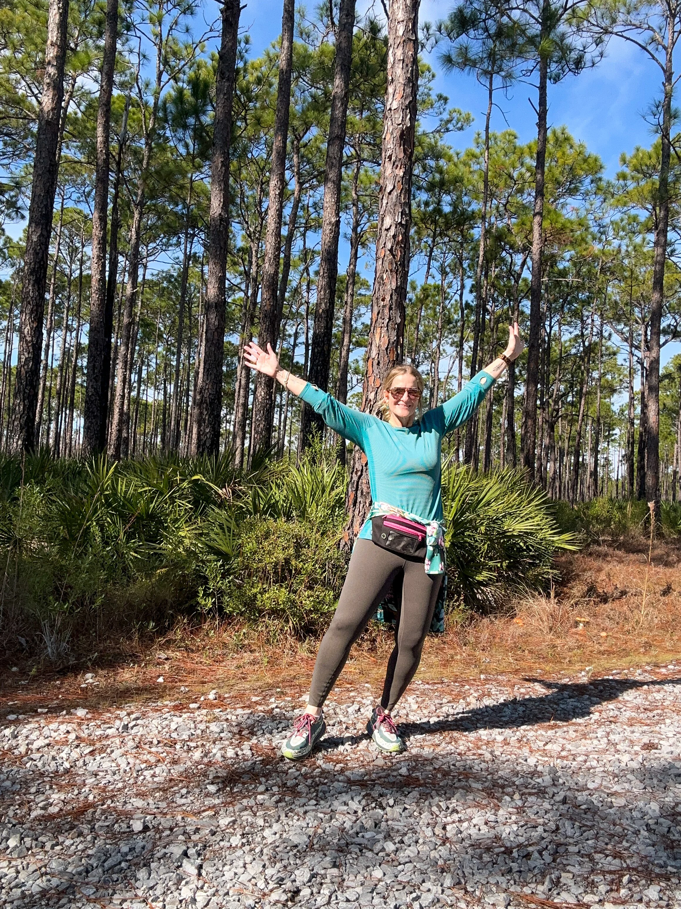 Pamela in hiking clothes, fanny pack and sunglasses with arms stretched in the air on a pine forest trail