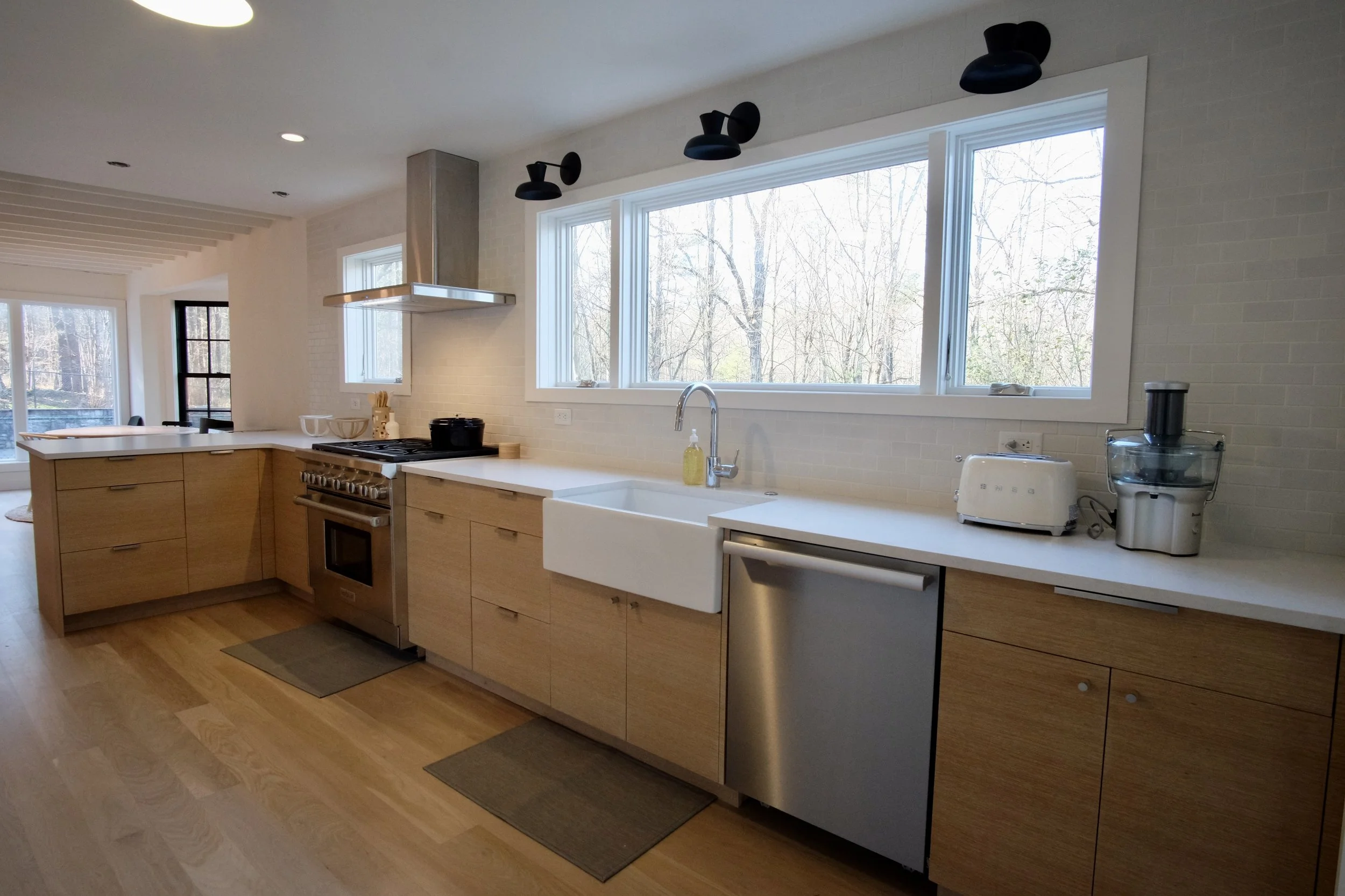 Modern kitchen with wooden cabinets, white countertops, a large window above the sink, and appliances including a stove, dishwasher, toaster, and juicer.