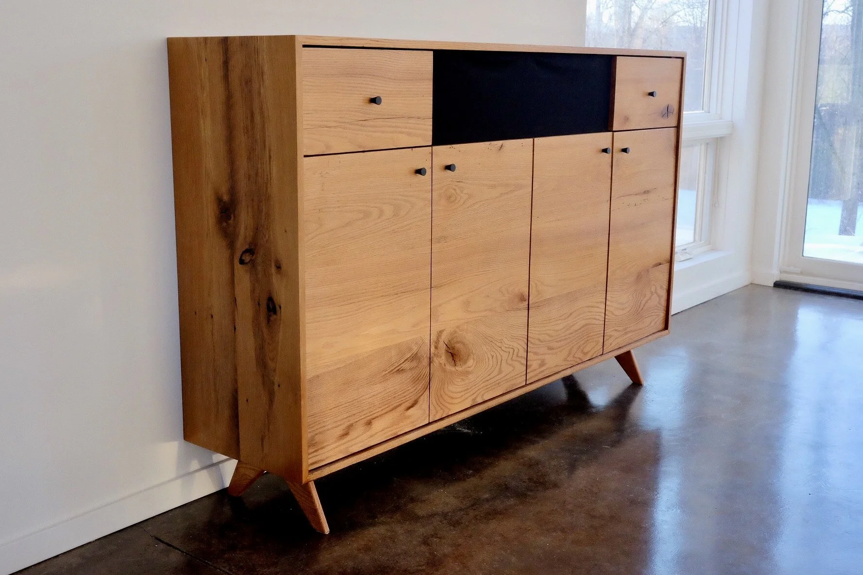 Mid-century modern wooden cabinet with black knobs, placed against a white wall near large windows, on a polished concrete floor.