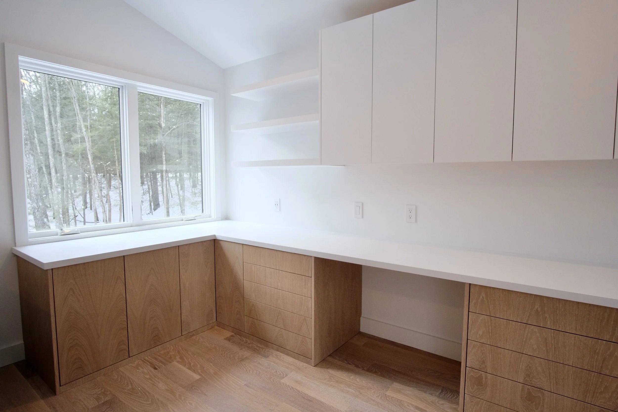 Modern kitchen with white countertops, wooden cabinets, and a large window overlooking a wooded area.