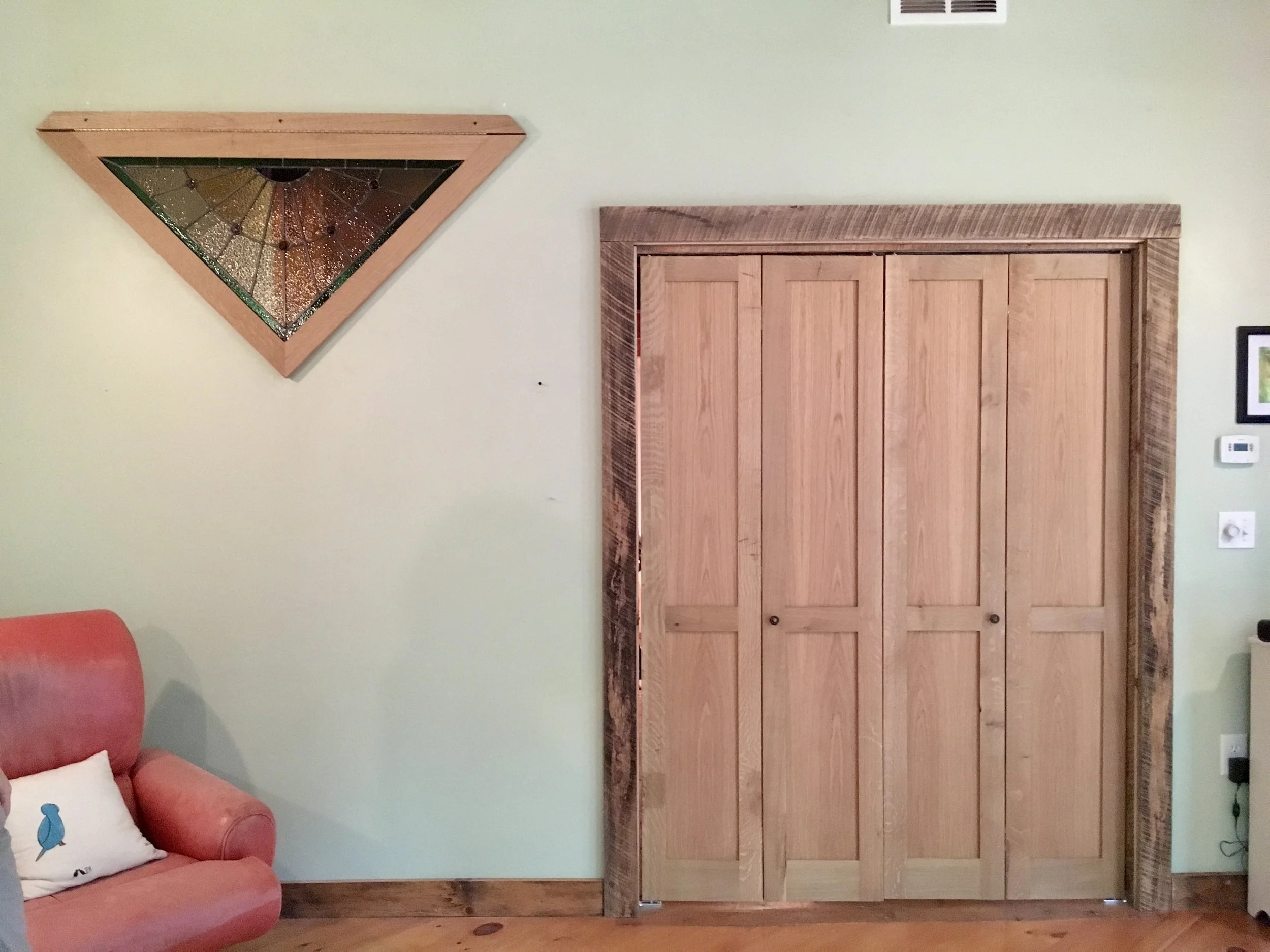 Living room wall featuring a closed wood closet with sliding doors, a decorative framed picture, and a wooden wall art piece resembling a stained glass window; part of a pink couch with an owl pillow is visible.
