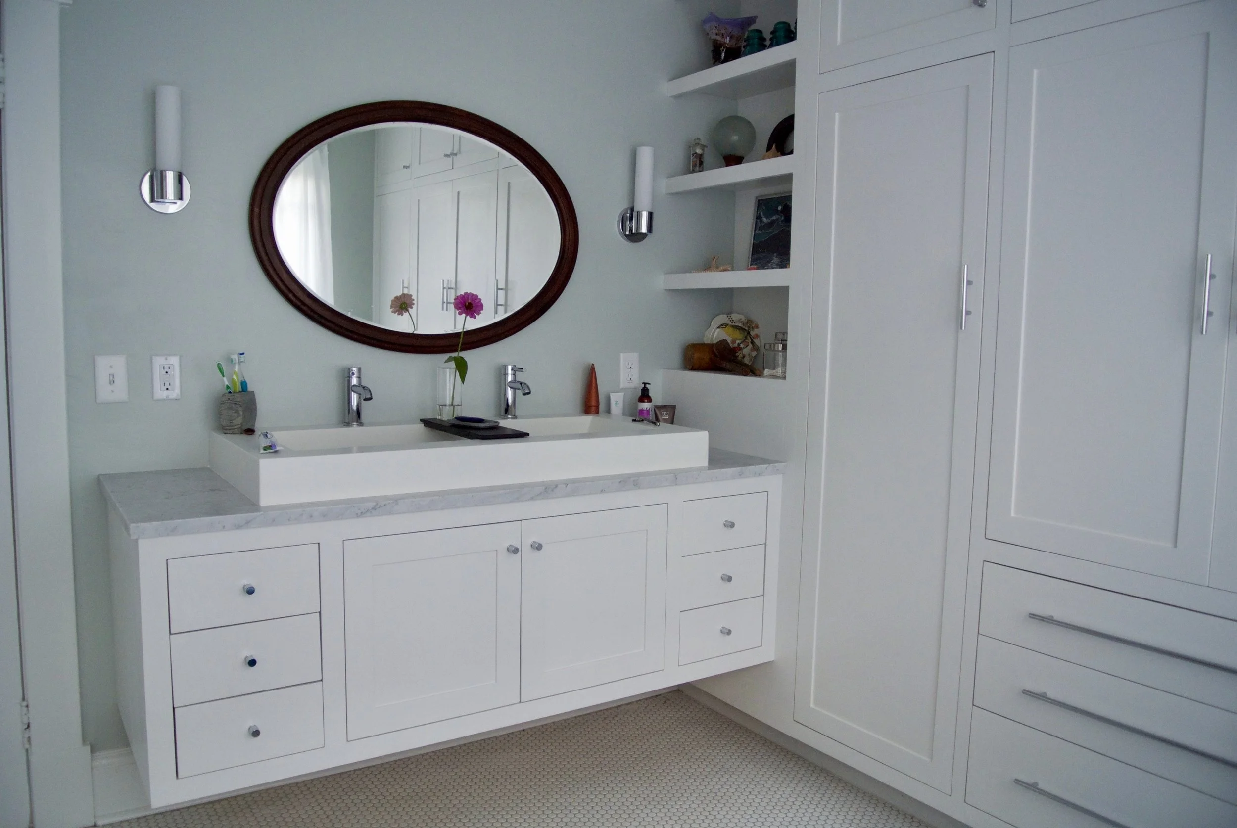 A white bathroom vanity with a marble countertop, a rectangular vessel sink, and a large oval mirror above it. The vanity has several drawers and cabinets, with two faucets and soap dispensers. There are two wall-mounted light fixtures and a vase wit
