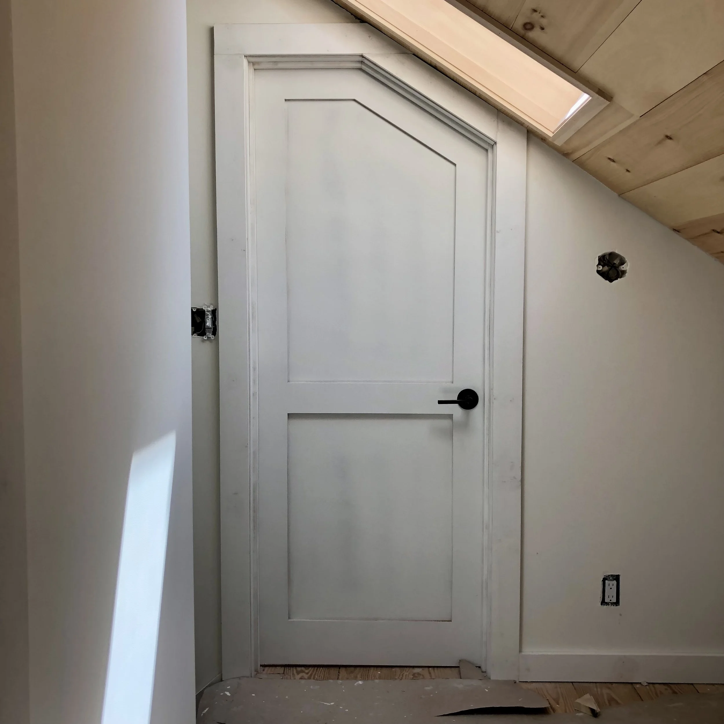 Interior wall with a white door and black handle, partially installed electrical outlet, and a ceiling with natural wood panels and a skylight.