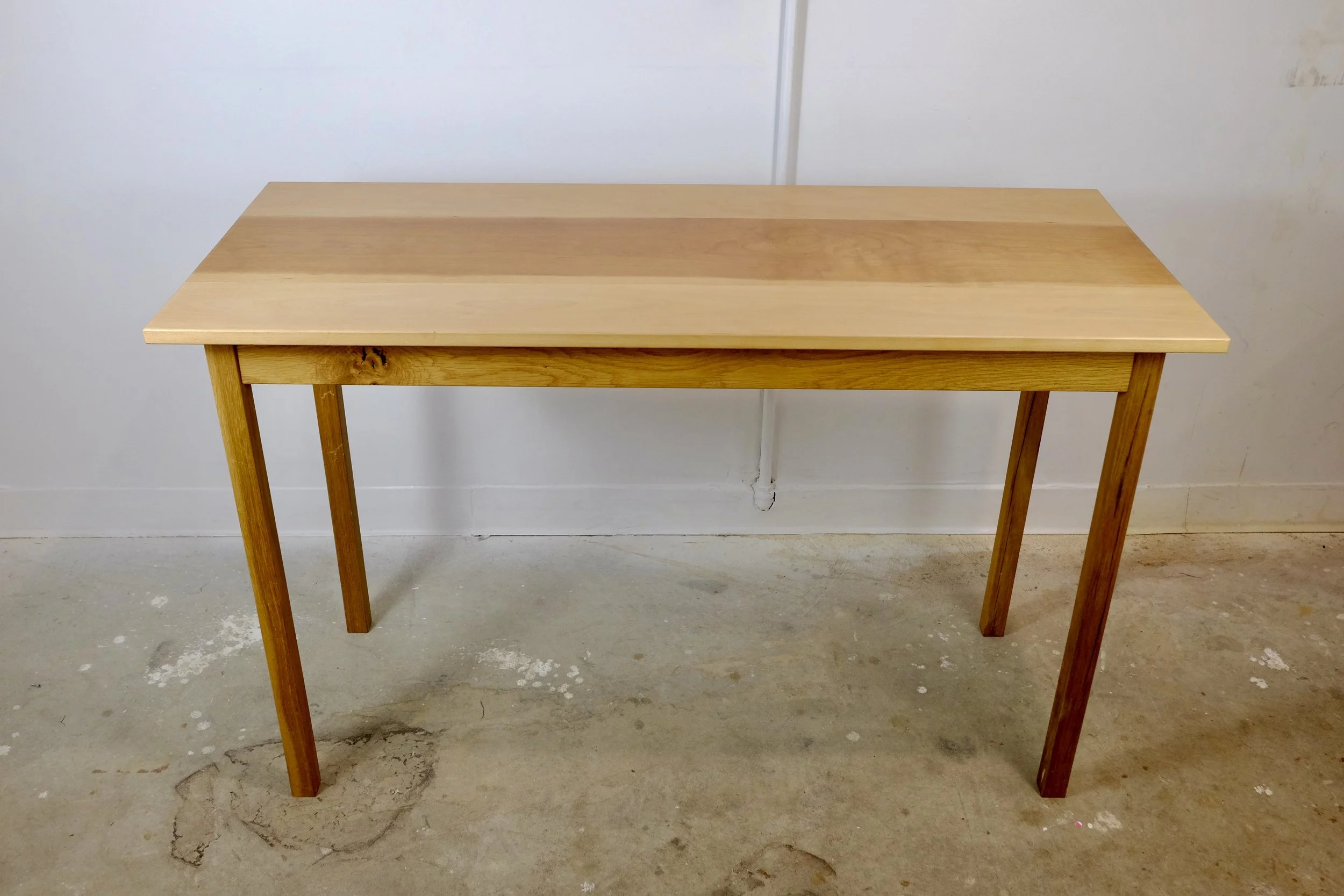 A wooden table with four legs, made of light-colored wood, placed on a concrete floor against a white wall background.