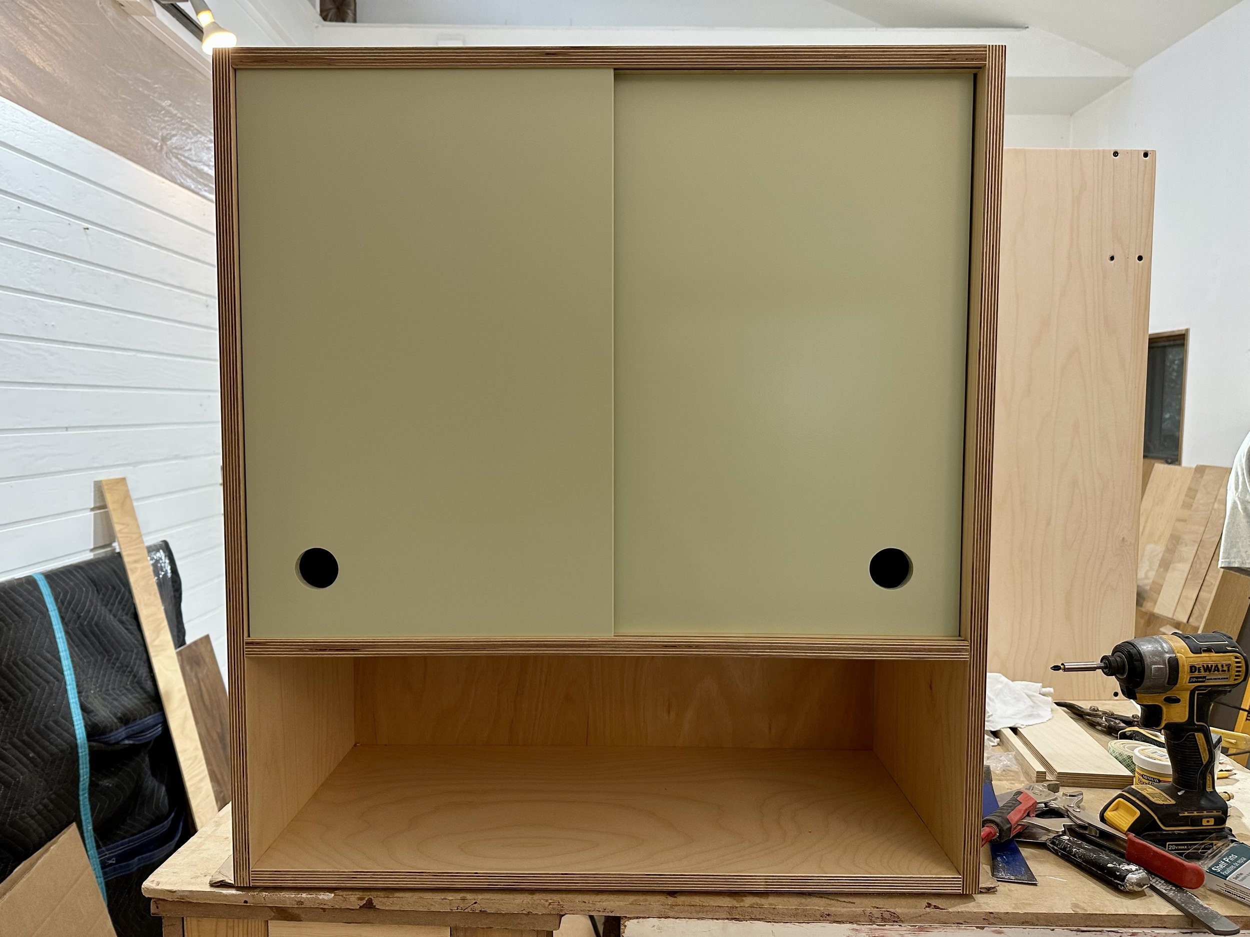 Wooden cabinet with sliding doors, partially open, in a workshop. Power drill and tools on a workbench nearby.