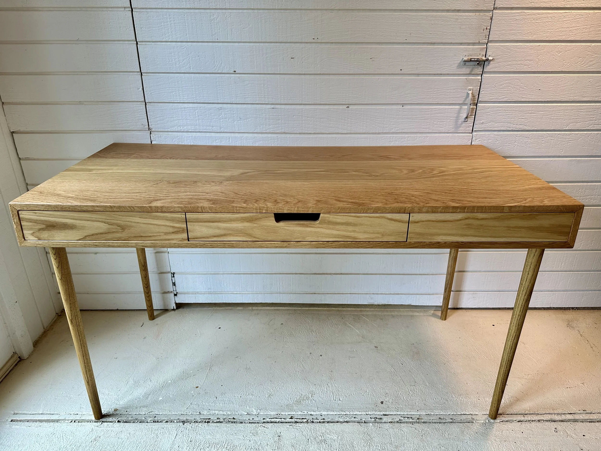A wooden table with three drawers and four angled legs, placed against a white wooden wall.