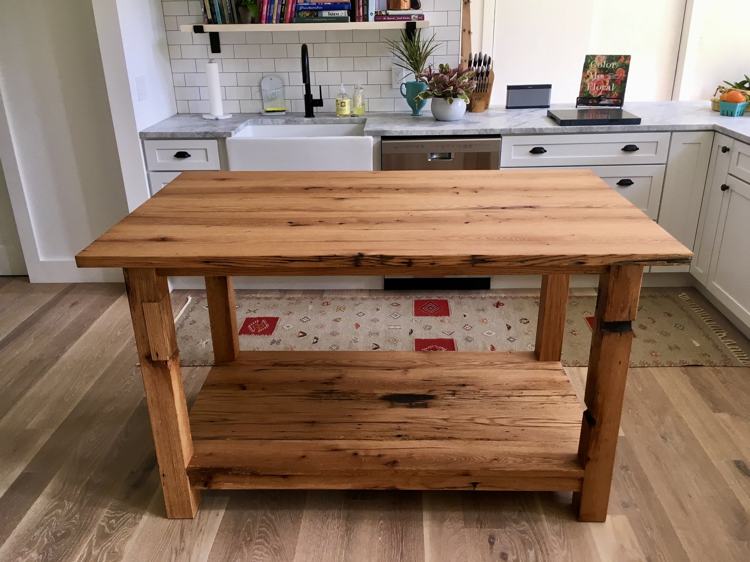 A wooden kitchen island with a butcher block top and lower shelf, in a kitchen with white cabinetry, a white sink, and various kitchen items on the countertop.