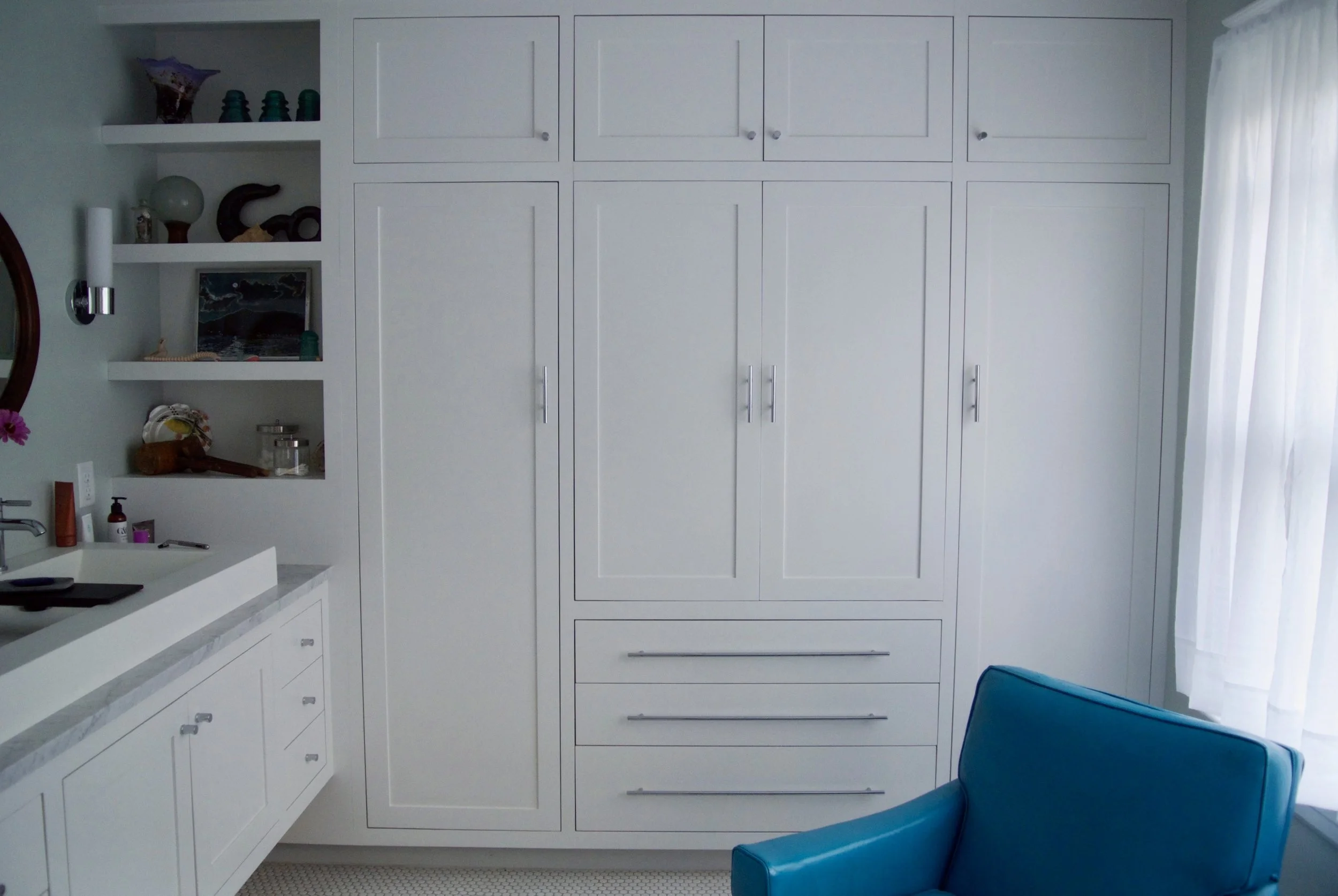 A white built-in closet with multiple doors and three drawers below, situated in a room with light-colored walls and sheer white curtains on a window to the right. Partially visible blue chair is in the foreground, and there are open shelves with dec