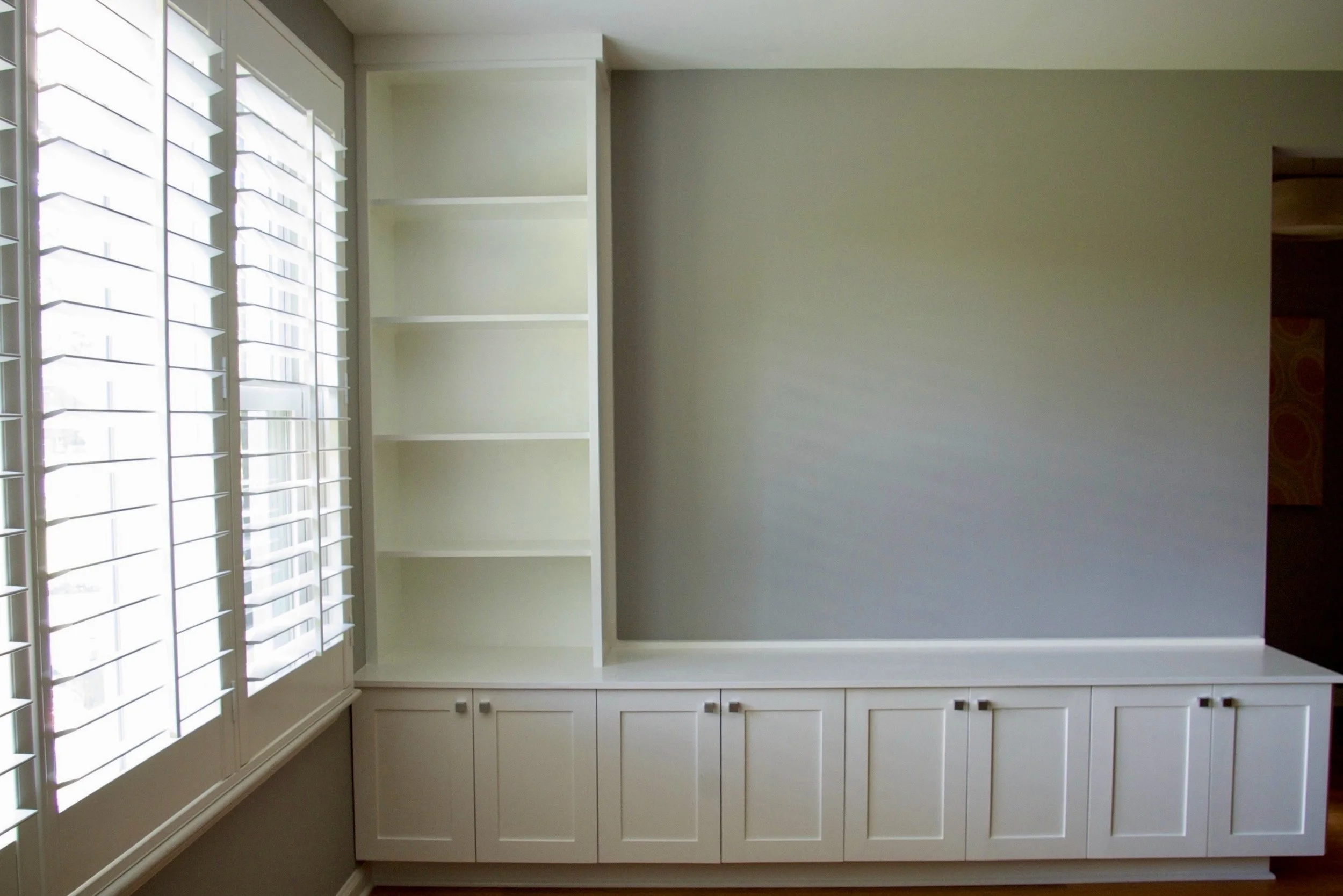 Empty white built-in cabinet with closed doors below and open shelves above, next to a window with white shutters, in a room with light-colored walls.