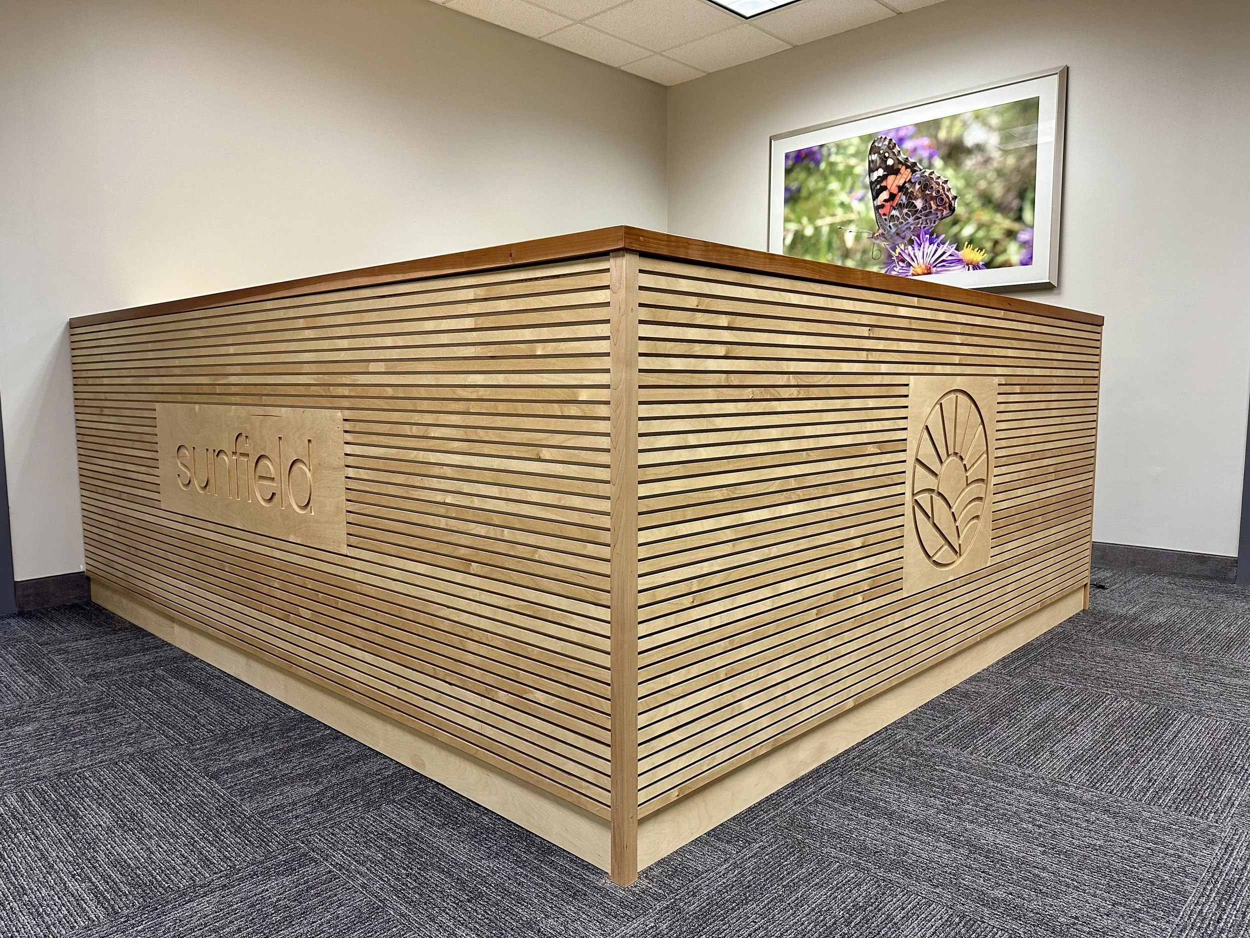 Wooden reception desk with the logo and name 'Sunfield' engraved on the side, in a modern office with a butterfly-exhibit picture on the wall.