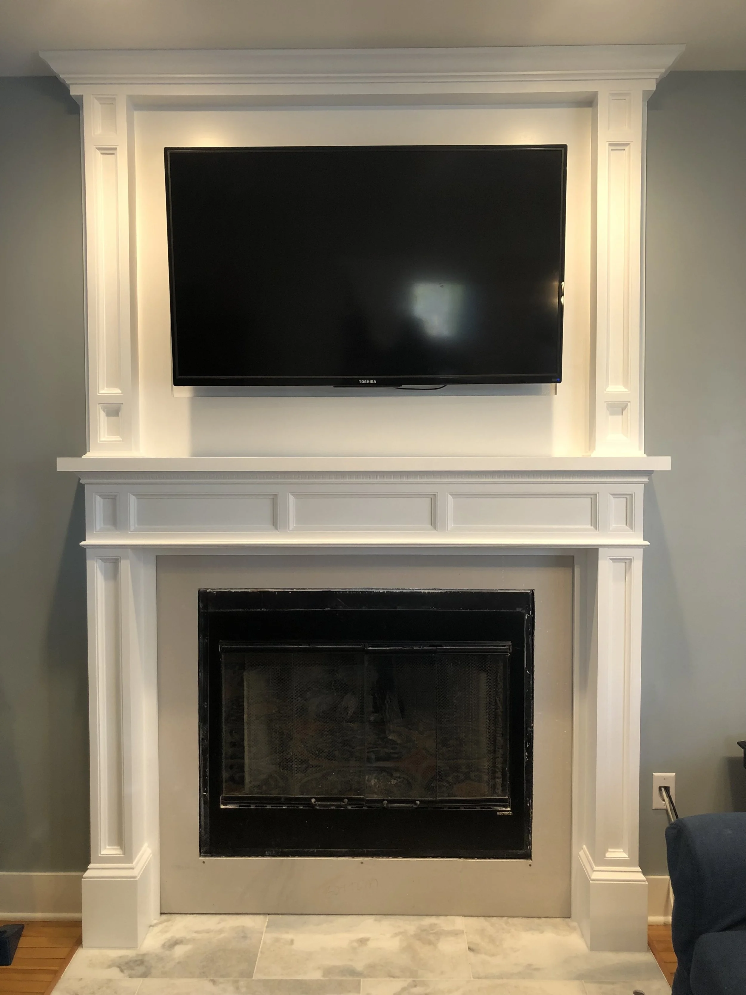 White fireplace with decorative molding and a mounted flat-screen TV above the mantel, set against a gray wall in a living room.
