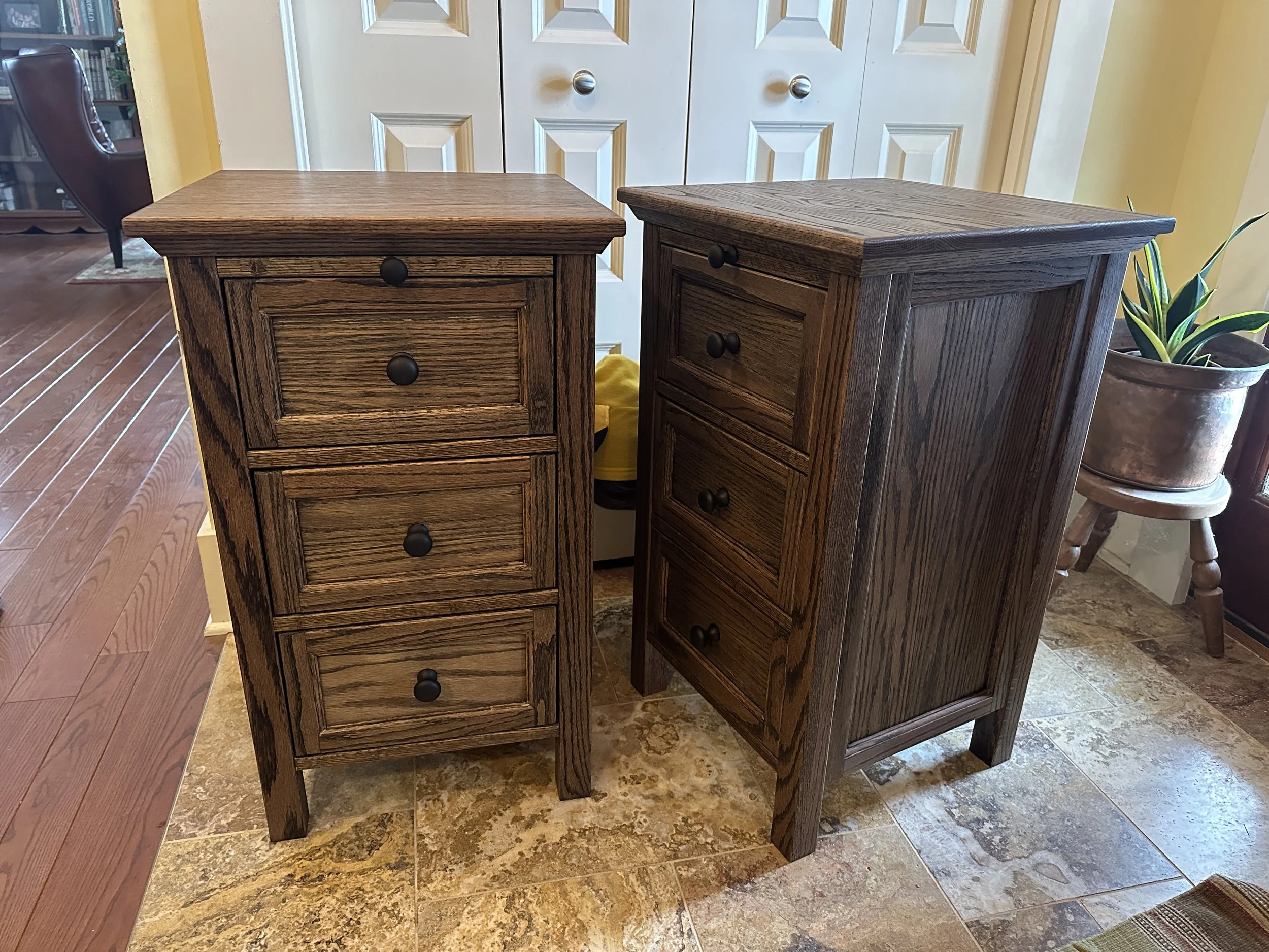 Two wooden bedside tables with three drawers each, placed next to each other on a tiled floor in a room.