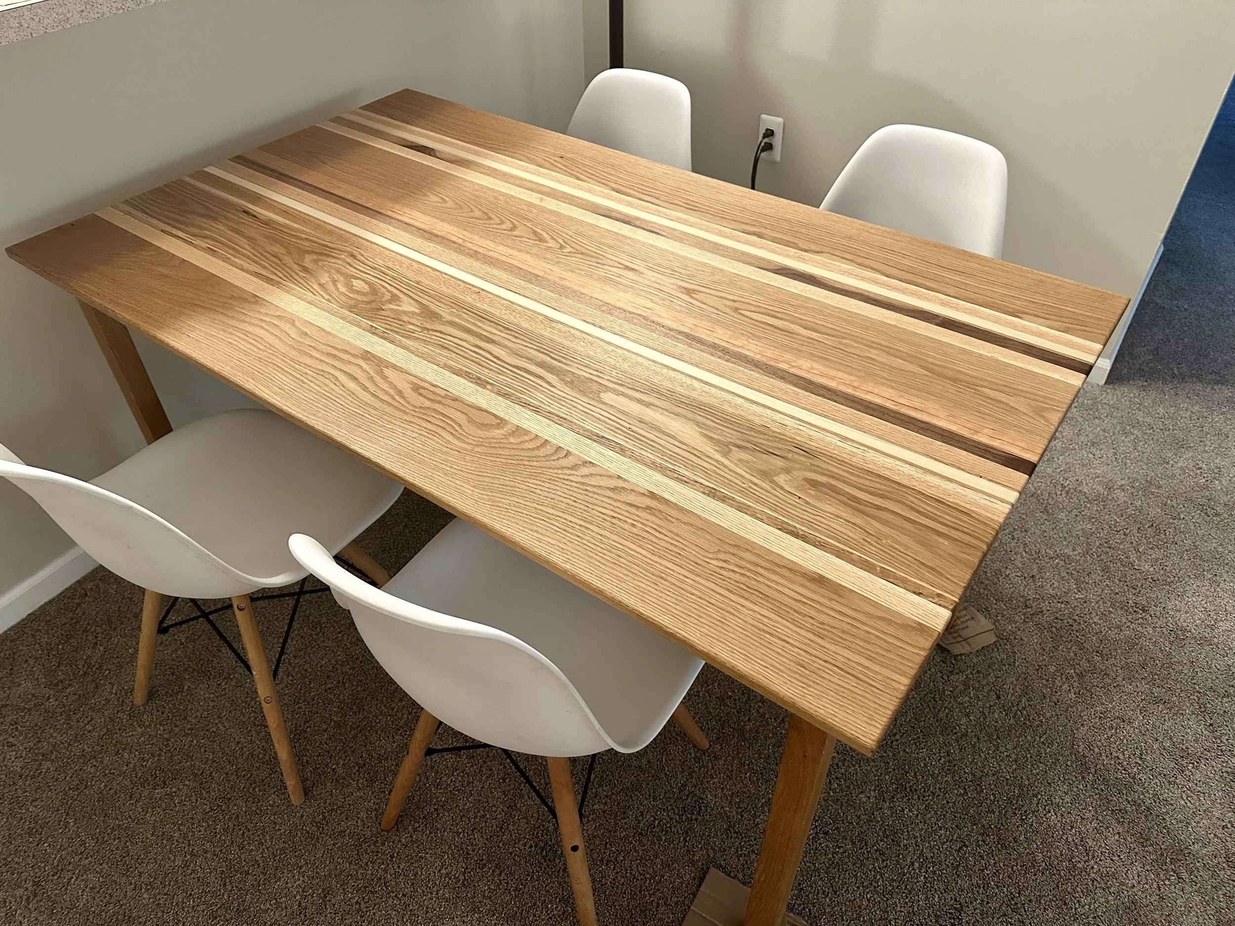 A wooden dining table with four white chairs around it in a room with beige carpet and white walls.